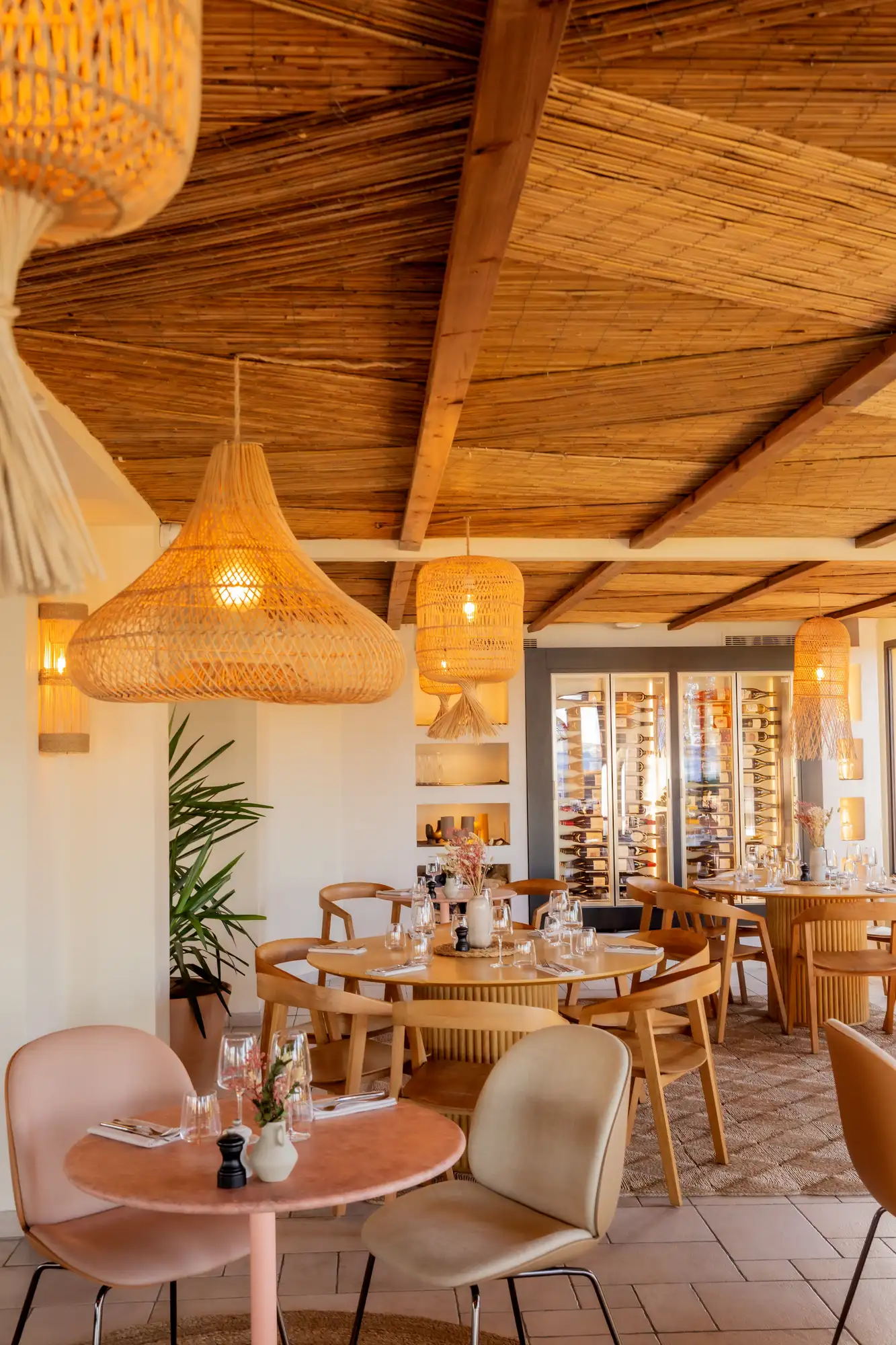 Intérieur chaleureux d'un restaurant avec des tables dressées, chaises modernes, suspensions en osier et étagère à bouteilles de vin en arrière-plan.