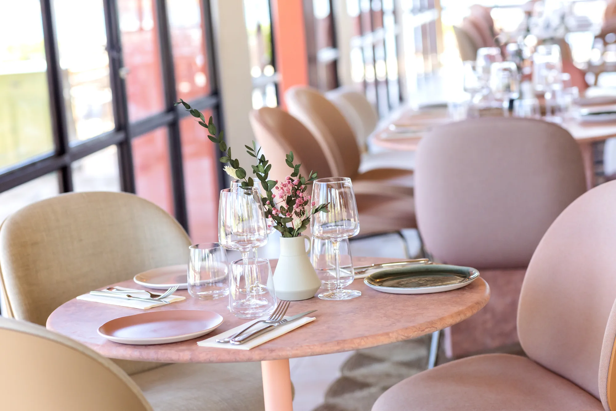 Table ronde dans un restaurant avec des chaises rembourrées roses et beige, décorée avec un vase blanc contenant des branches vertes et des petites fleurs roses, assiettes, couverts, et verres à vin et à eau.