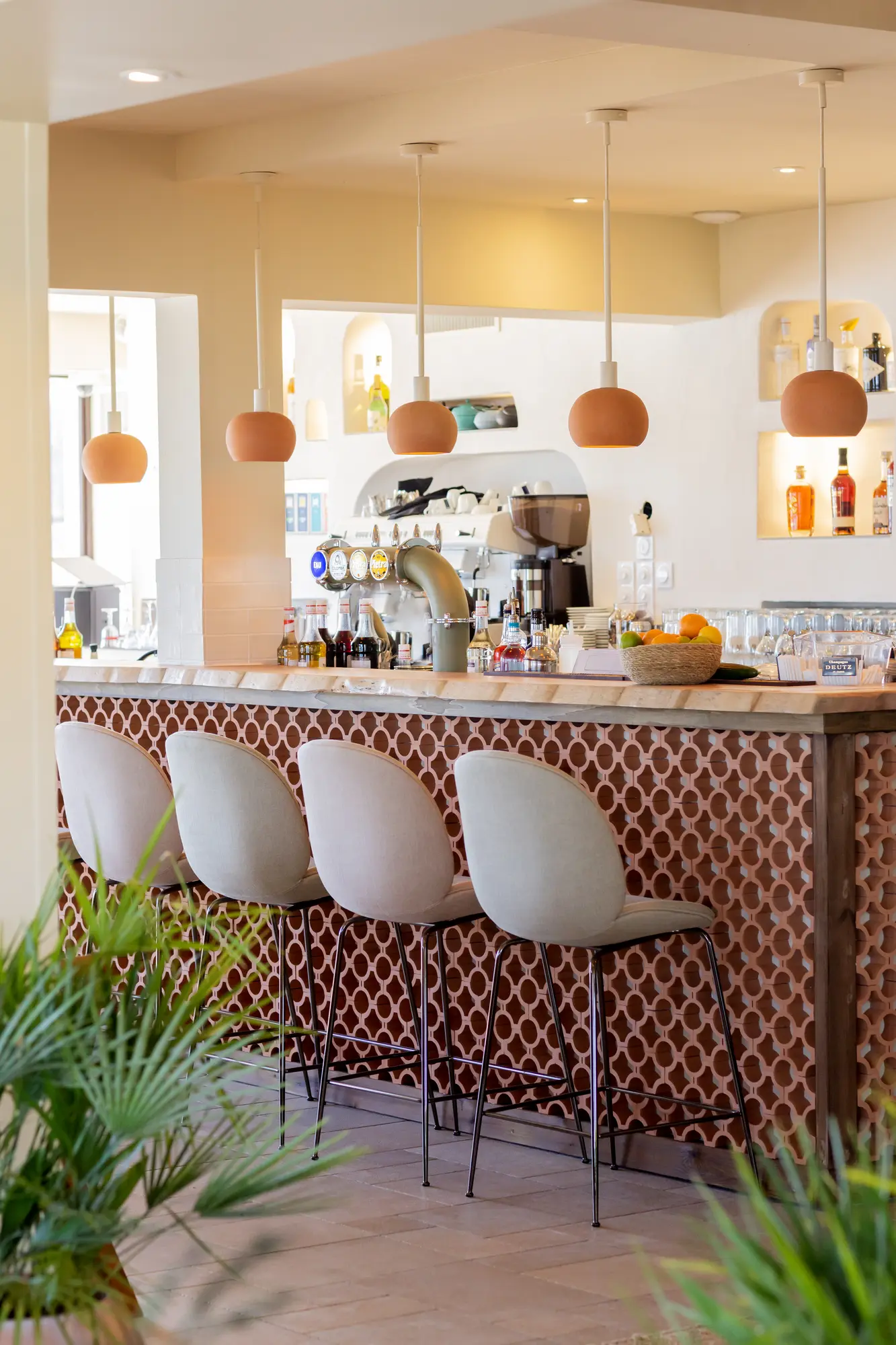 Coin bar moderne avec quatre chaises hautes beige devant un comptoir en bois à motifs géométriques marron, suspensions rondes suspendues au plafond, et divers bouteilles et verres sur le comptoir dans un espace lumineux et épuré.