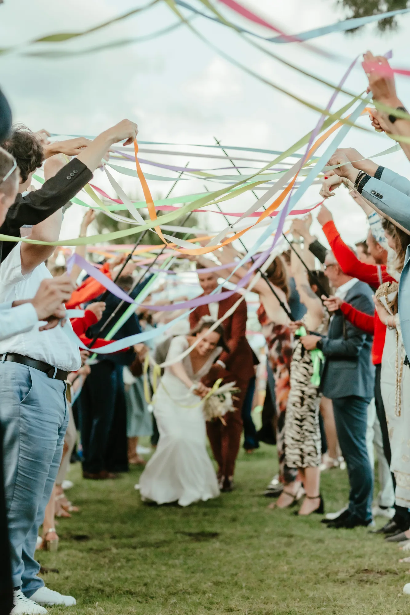 Des invités sur un chemin en plein air tiennent des rubans colorés levés, formant une voûte sous laquelle un couple de mariés souriants s'avance, la mariée en robe blanche tenant un bouquet.