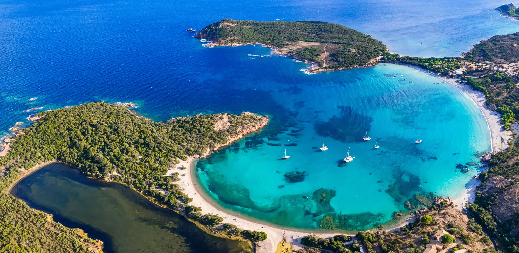 Vue aérienne d’une baie aux eaux turquoise avec plusieurs voiliers ancrés, une plage de sable clair et une île boisée adjacente entourée par la mer bleue profonde.