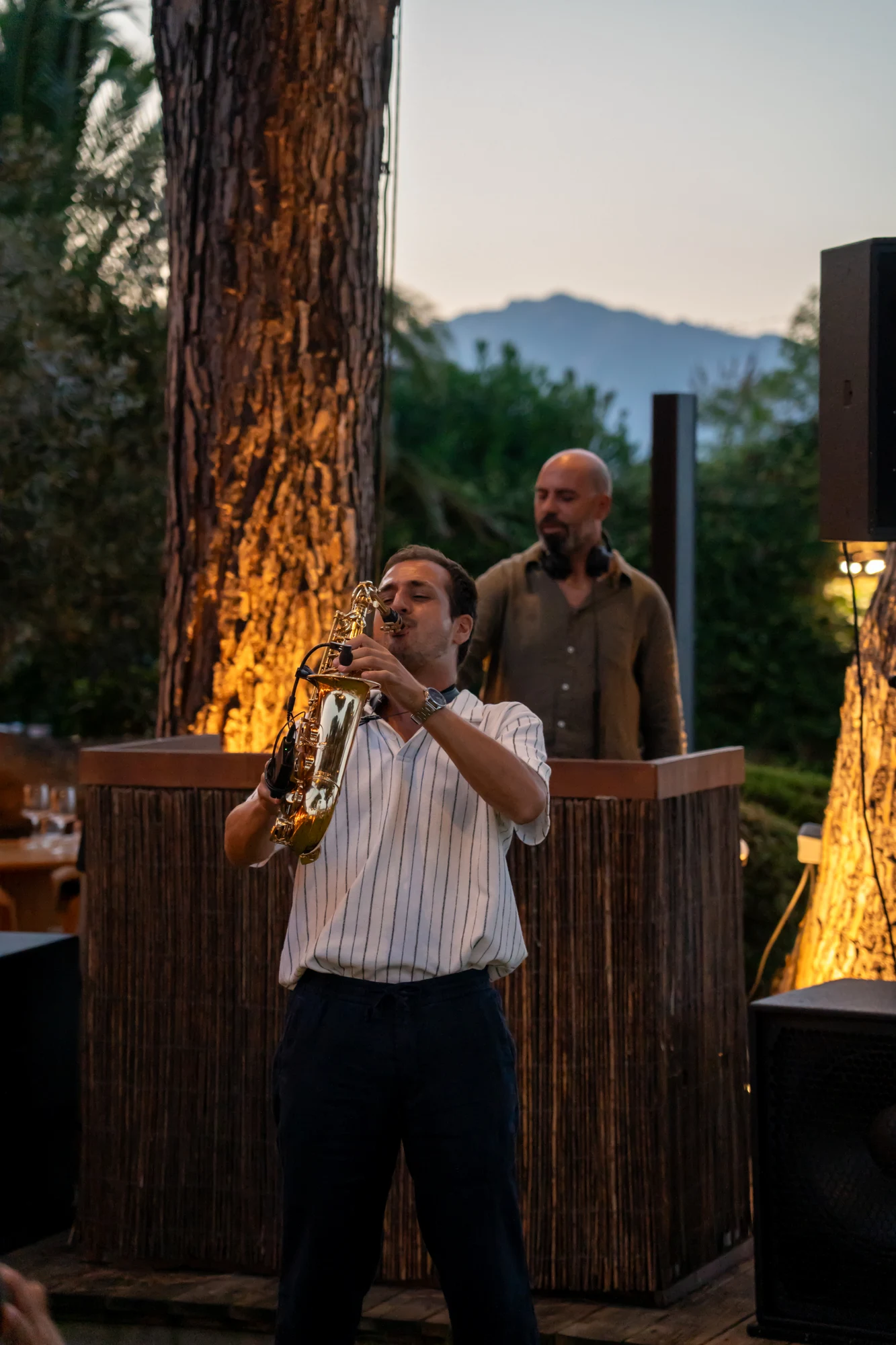 Musicien jouant du saxophone devant un DJ dans un cadre extérieur avec un grand arbre et des montagnes en arrière-plan au crépuscule.