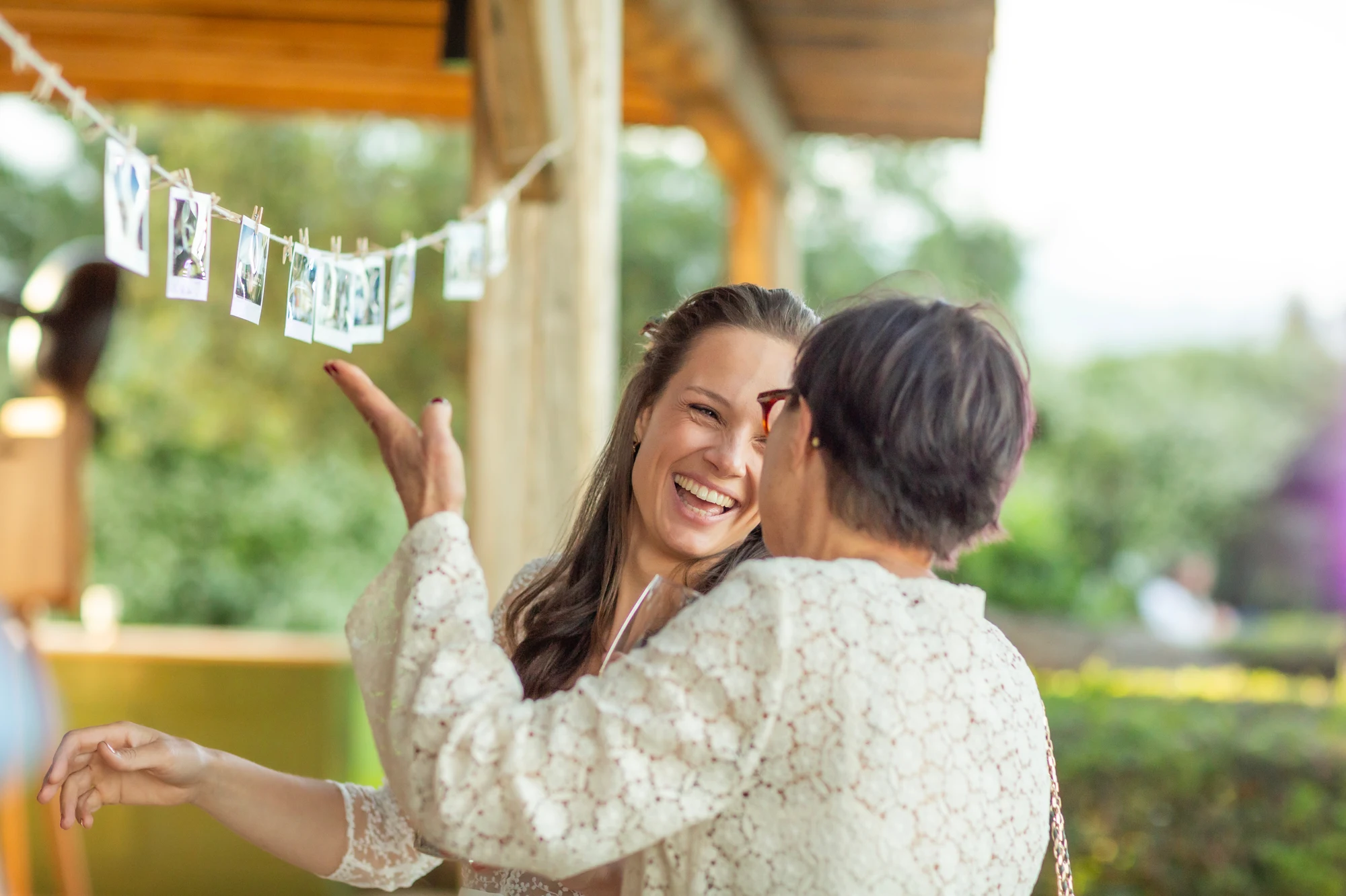 Deux femmes souriantes en tenue dentelle discutant joyeusement à une réception en plein air avec des photos suspendues en arrière-plan.