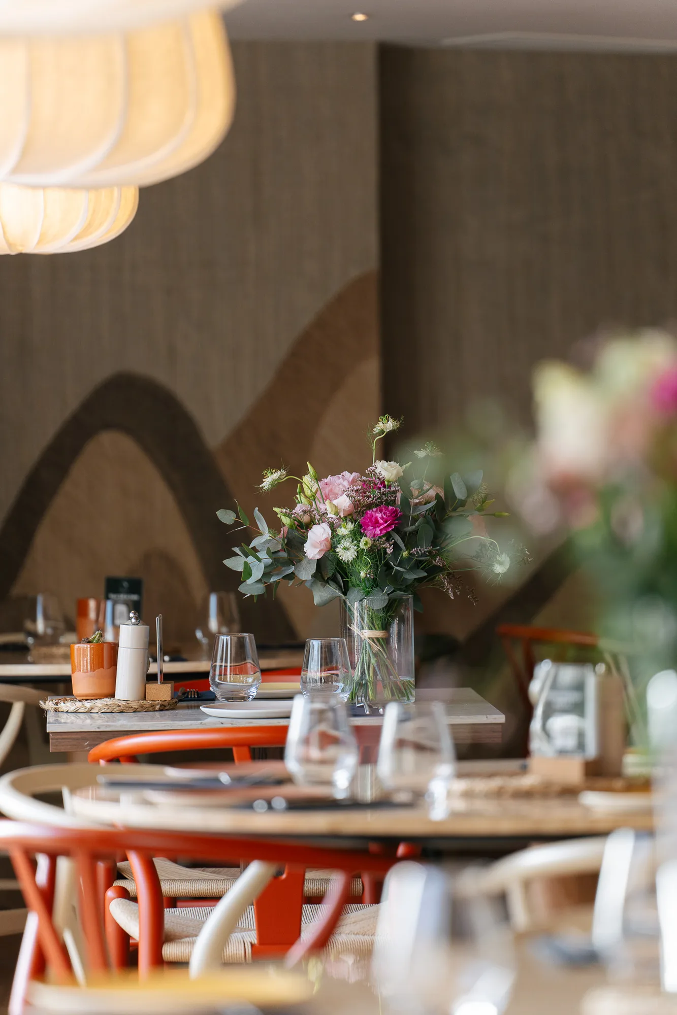 Intérieur d'un restaurant moderne avec des tables en bois, des chaises en bois claires et oranges, des verres à eau et une grande composition florale colorée au centre d'une table.