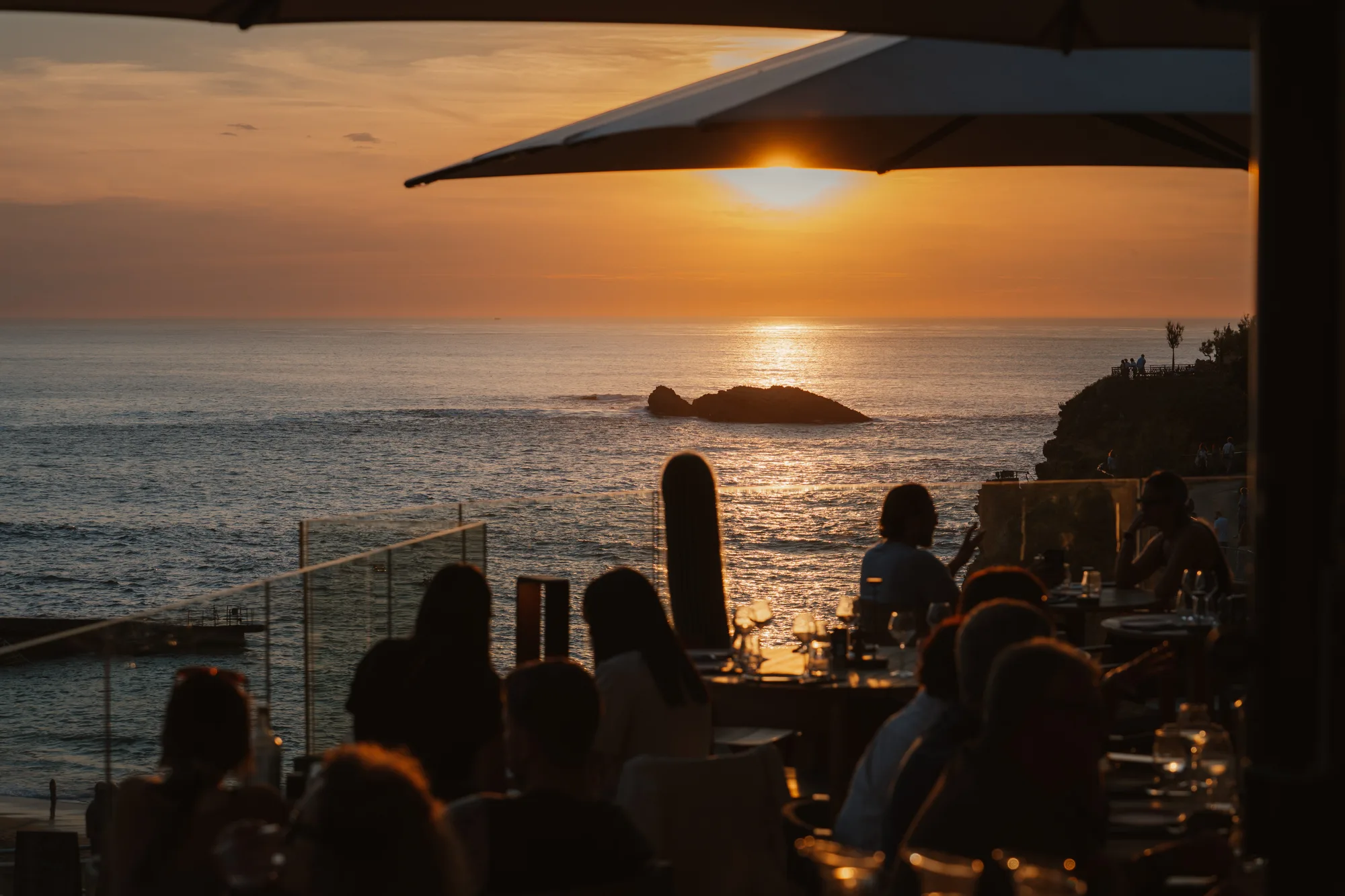 Coucher de soleil orange sur l'océan avec un rocher visible au loin, vu depuis une terrasse de restaurant où des silhouettes de personnes sont assises à des tables sous un grand parasol.