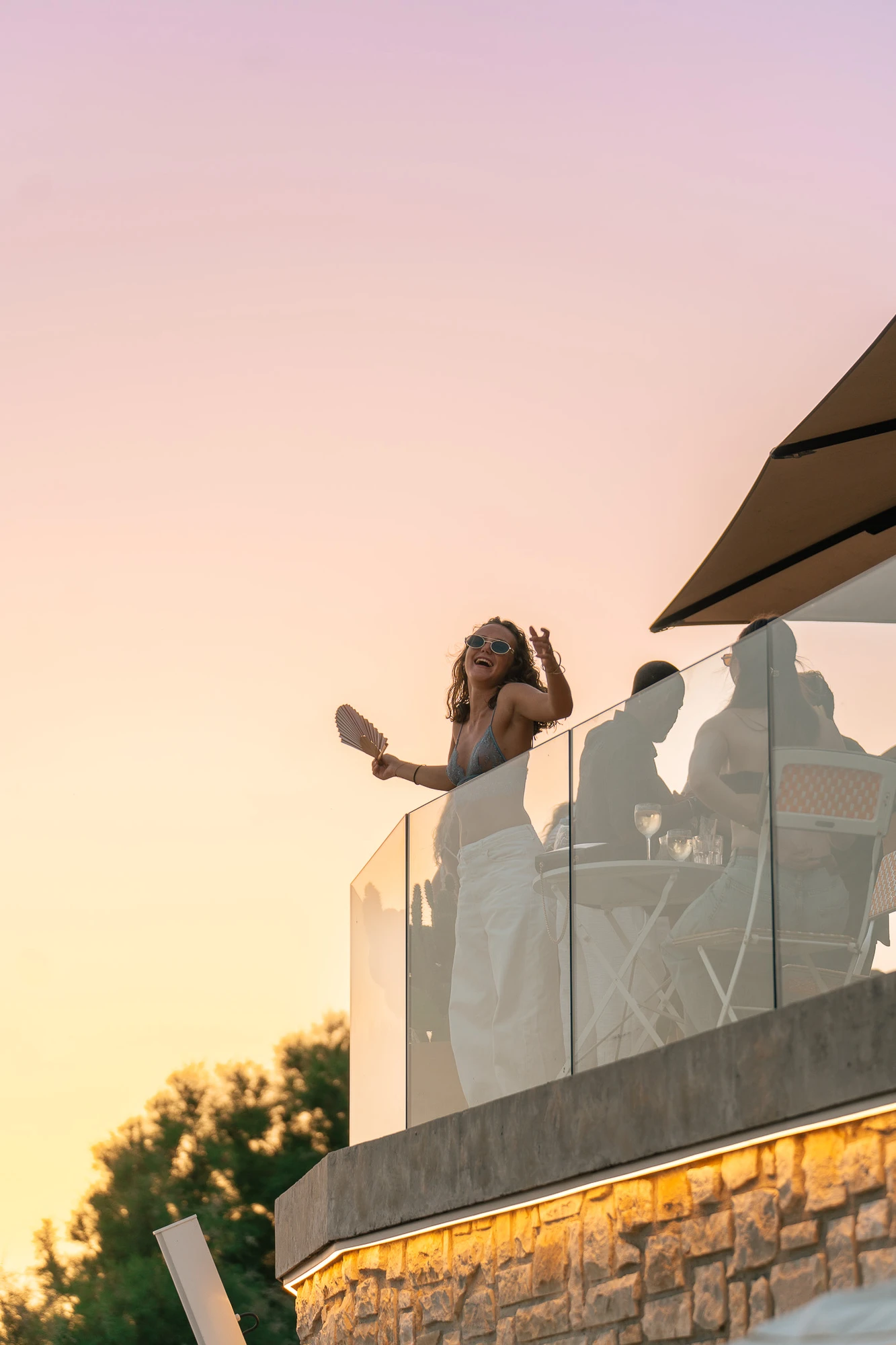 Jeune femme souriante portant des lunettes de soleil et un haut léger, se tenant sur un balcon en verre au coucher du soleil, tenant un éventail et faisant un geste de la main, avec des personnes assises à une table derrière elle.