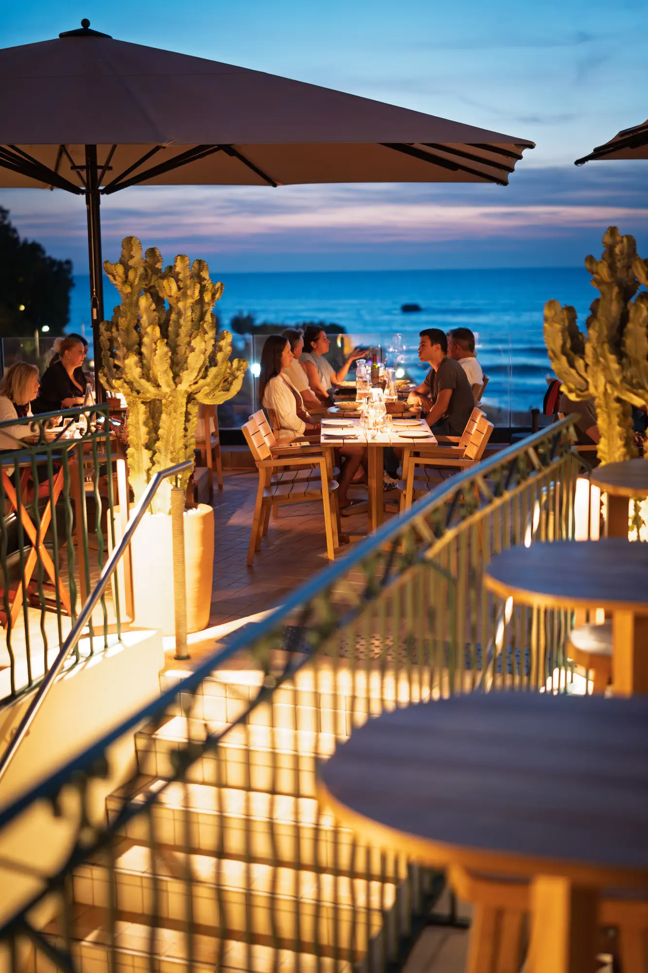 Terrasse de restaurant éclairée en soirée avec des clients assis autour d'une table en bois sous un grand parasol, vue sur l'océan au coucher du soleil et cactus décoratifs.