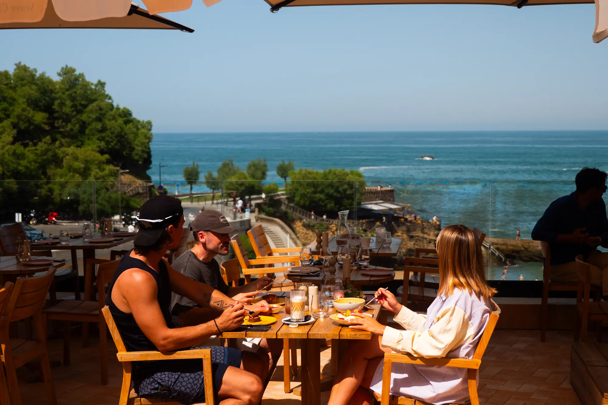Trois personnes assises autour d'une table en bois sur une terrasse avec vue sur la mer, mangeant et discutant sous un parasol.