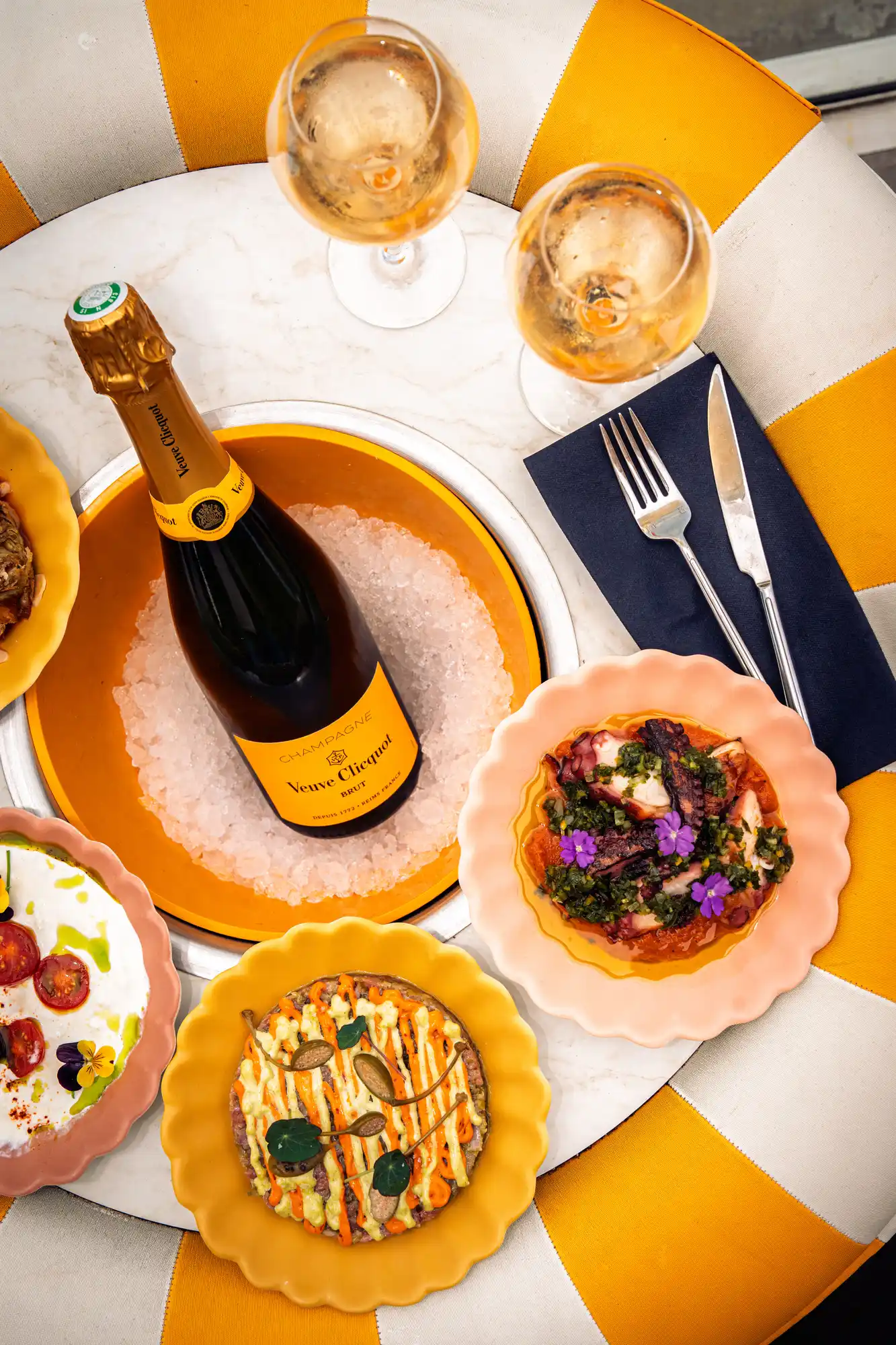 Table ronde avec une bouteille de champagne Veuve Clicquot sur glace au centre, deux verres de champagne, plusieurs assiettes colorées avec des plats gastronomiques et des couverts posés sur une serviette noire, le tout sur une chaise rayée jaune et blanche.