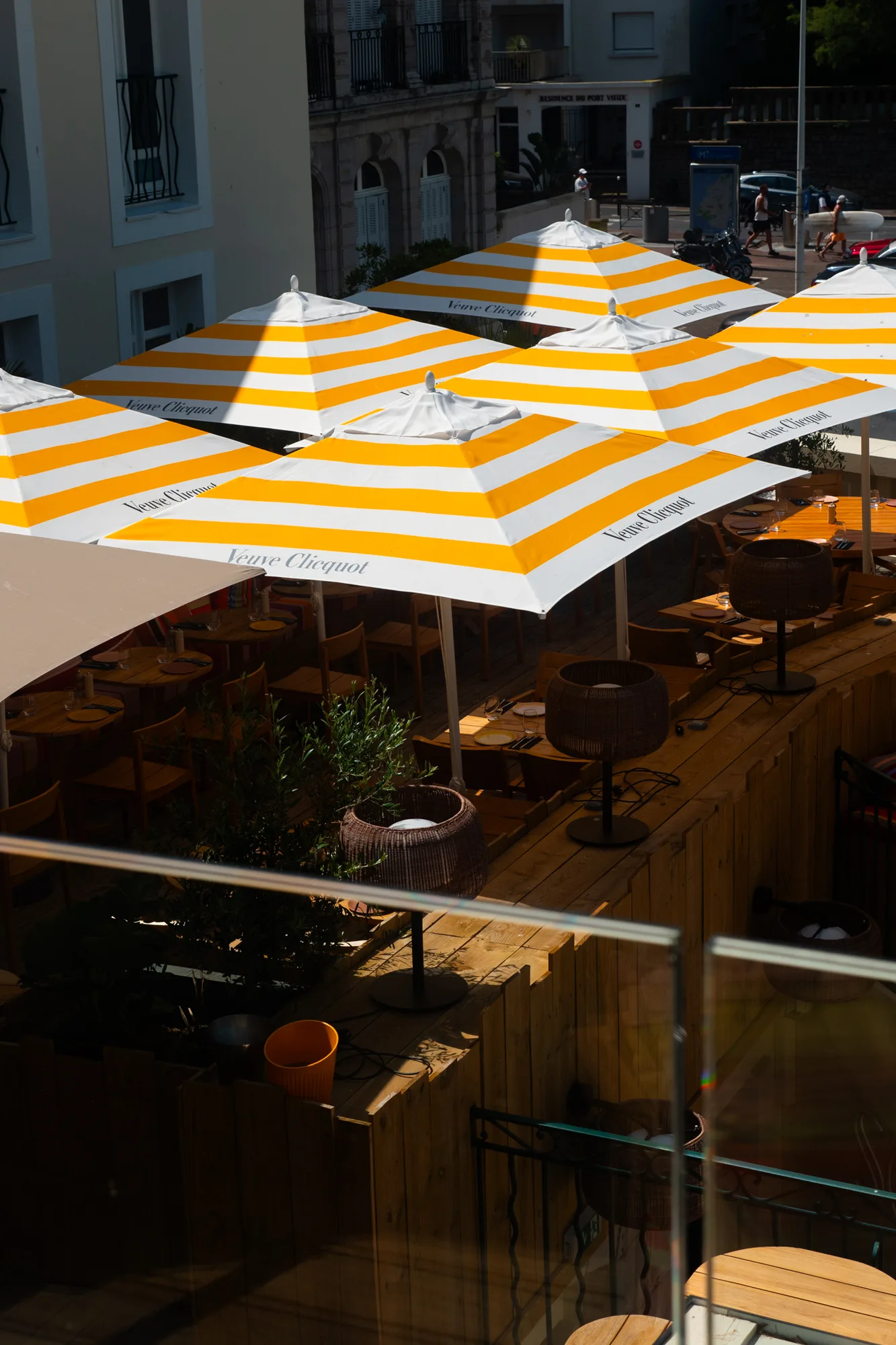 Terrasse de café extérieure avec plusieurs parasols à rayures blanches et jaunes portant le logo Veuve Clicquot, tables en bois dressées avec vaisselle et verres, éclairage d'ambiance et décor végétal.