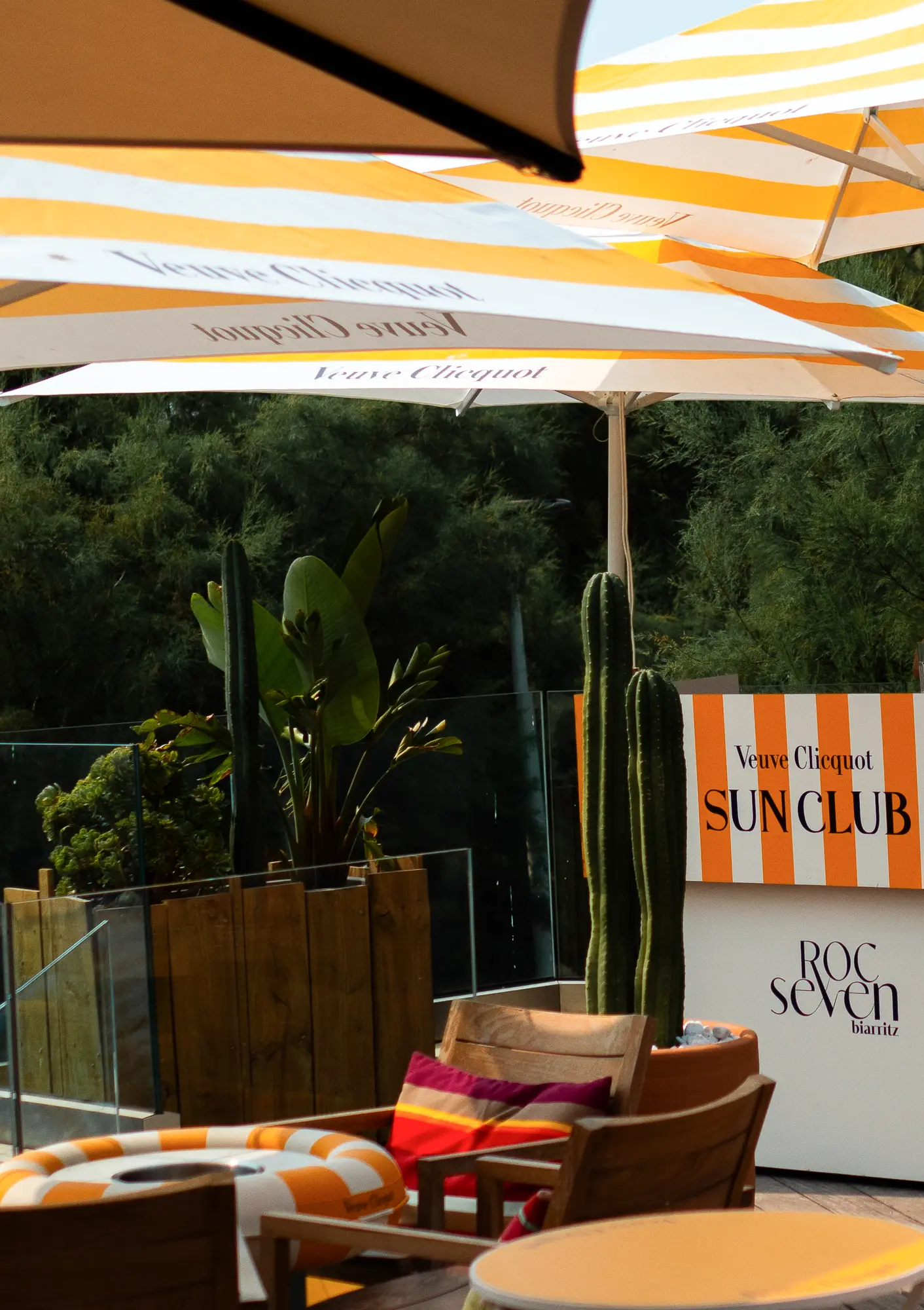 Terrasse extérieure avec parasols jaunes et blancs portant l'inscription Veuve Clicquot, fauteuils en bois avec coussins colorés, plantes dont des cactus, et une enseigne rayée orange et blanche indiquant Veuve Clicquot SUN CLUB et Roc Seven Biarritz.