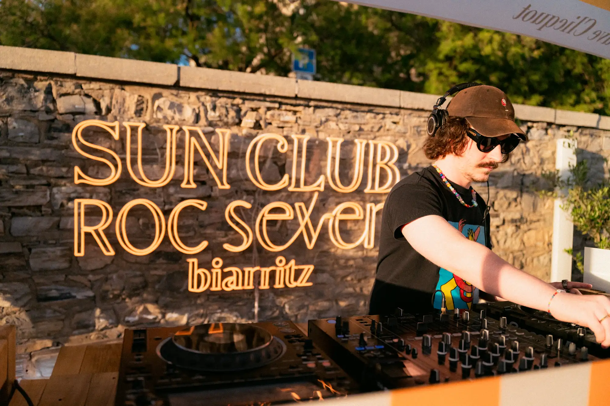 DJ portant une casquette, des lunettes de soleil, un collier coloré et un t-shirt noir, maniant une table de mixage extérieure devant un mur de pierre avec le texte lumineux 'SUN CLUB ROC seven biarritz'.