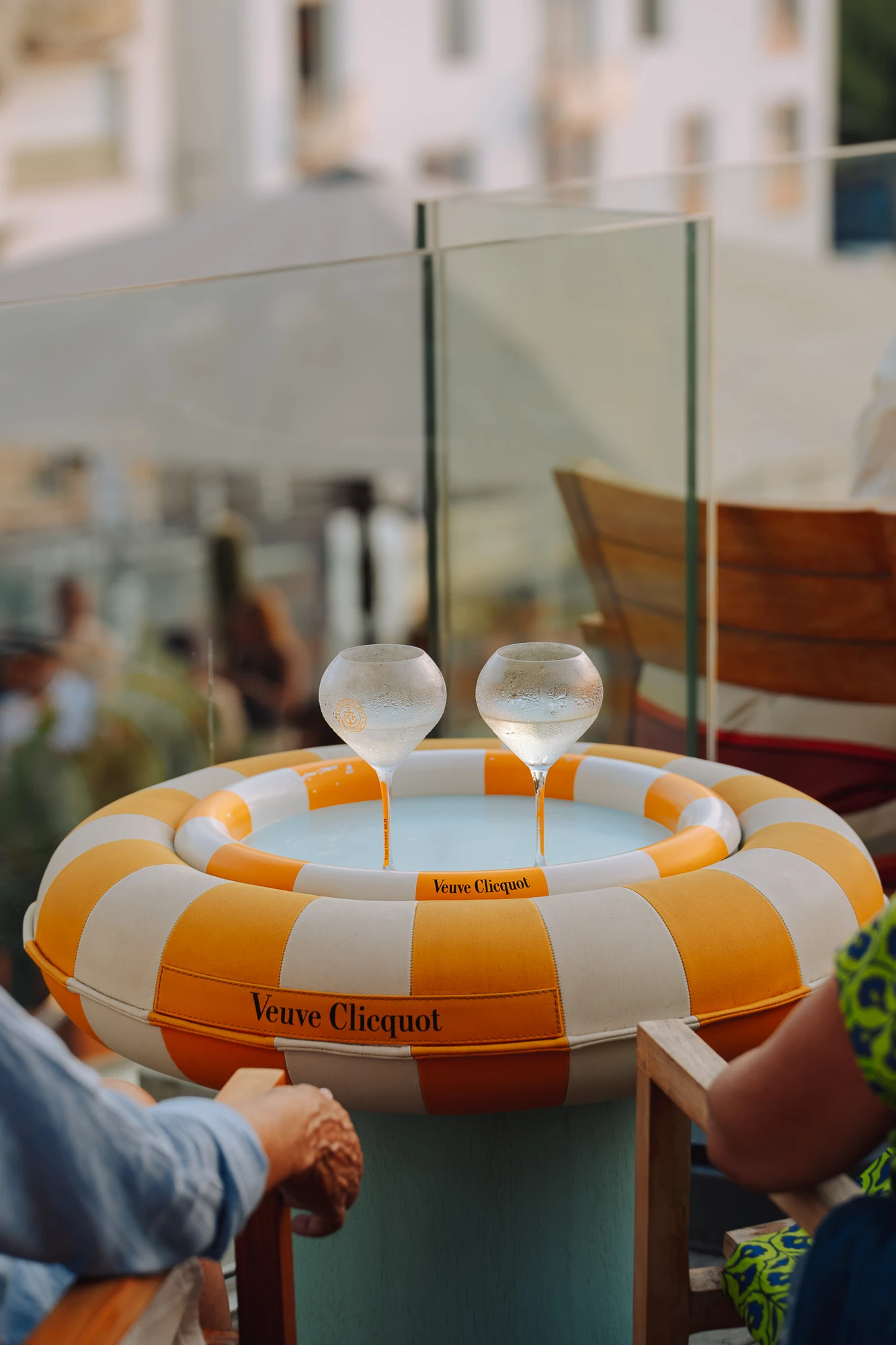 Deux verres à pied givrés remplis d'eau posés dans une bouée jaune et blanche rayée marquée Veuve Clicquot, utilisée comme support de table entre deux personnes assises.