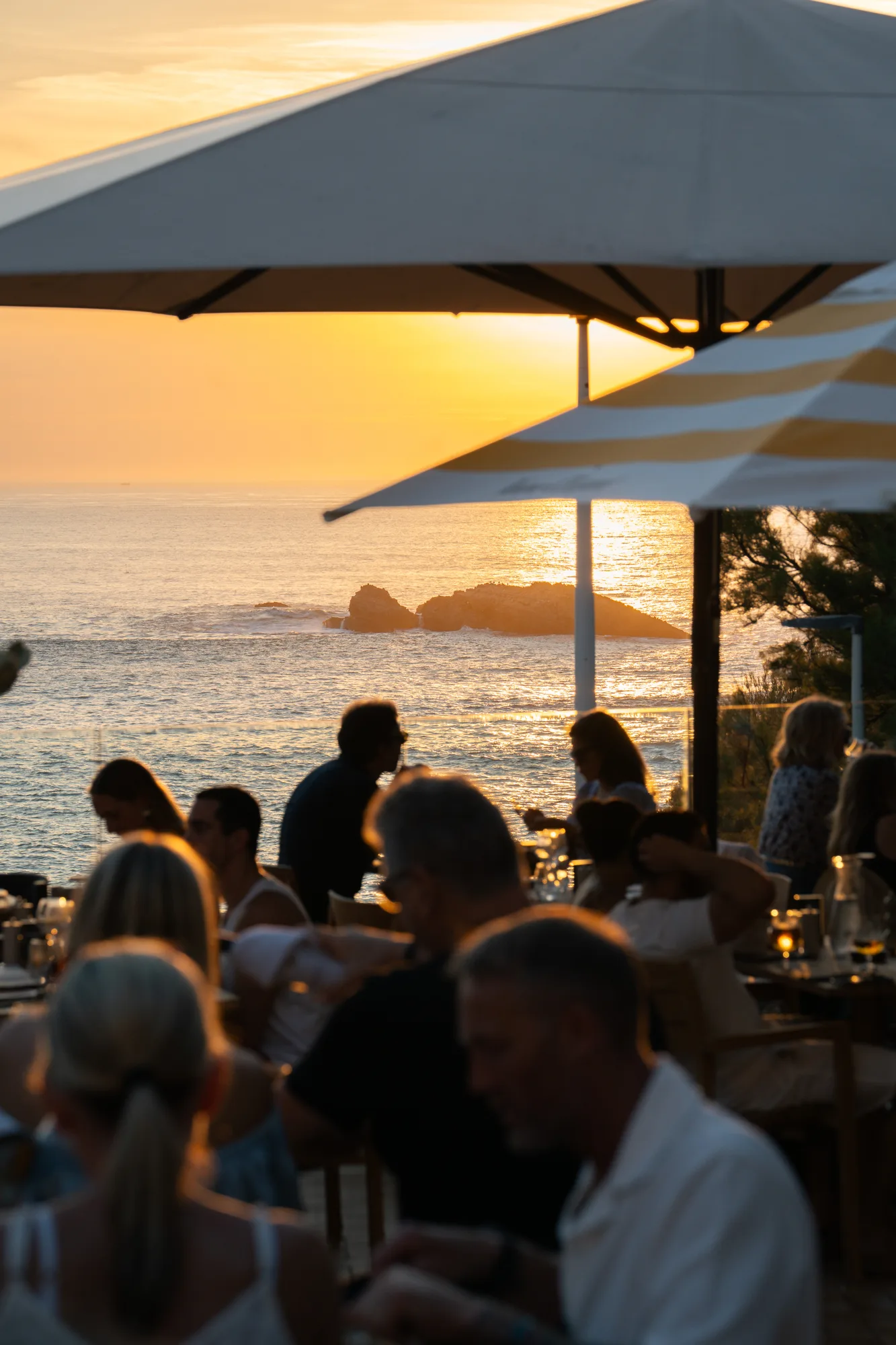 Personnes assises sous de grands parasols dans un restaurant en plein air au bord de la mer au coucher du soleil, avec une petite île rocheuse en arrière-plan enveloppée par la lumière dorée.