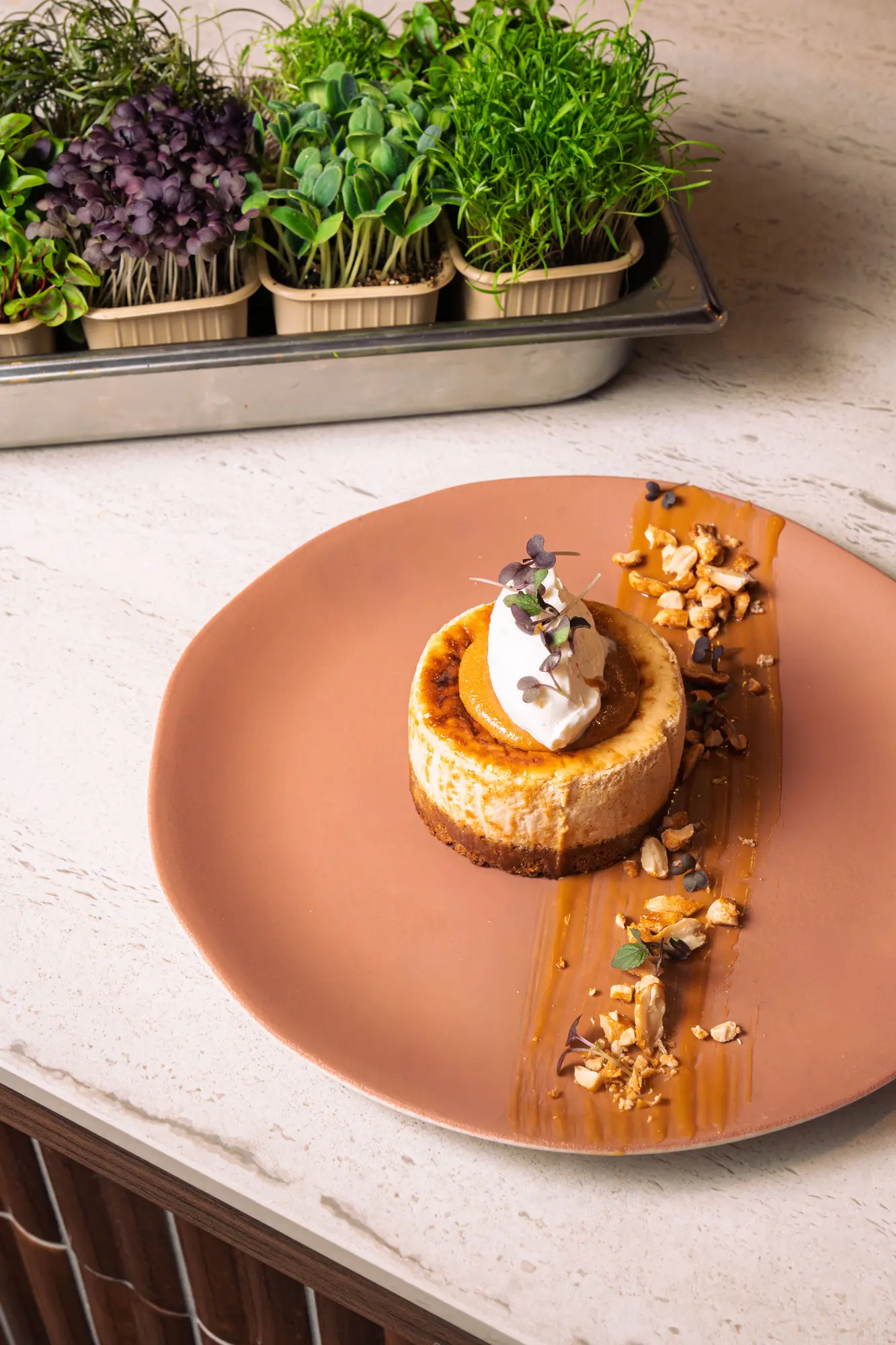 Un dessert gourmet rond sur une grande assiette marron, garni de crème fouettée, sauce caramel ou butterscotch, éclats de noix et micro-pousses, sur un comptoir clair avec un plateau de jeunes pousses vertes en arrière-plan.