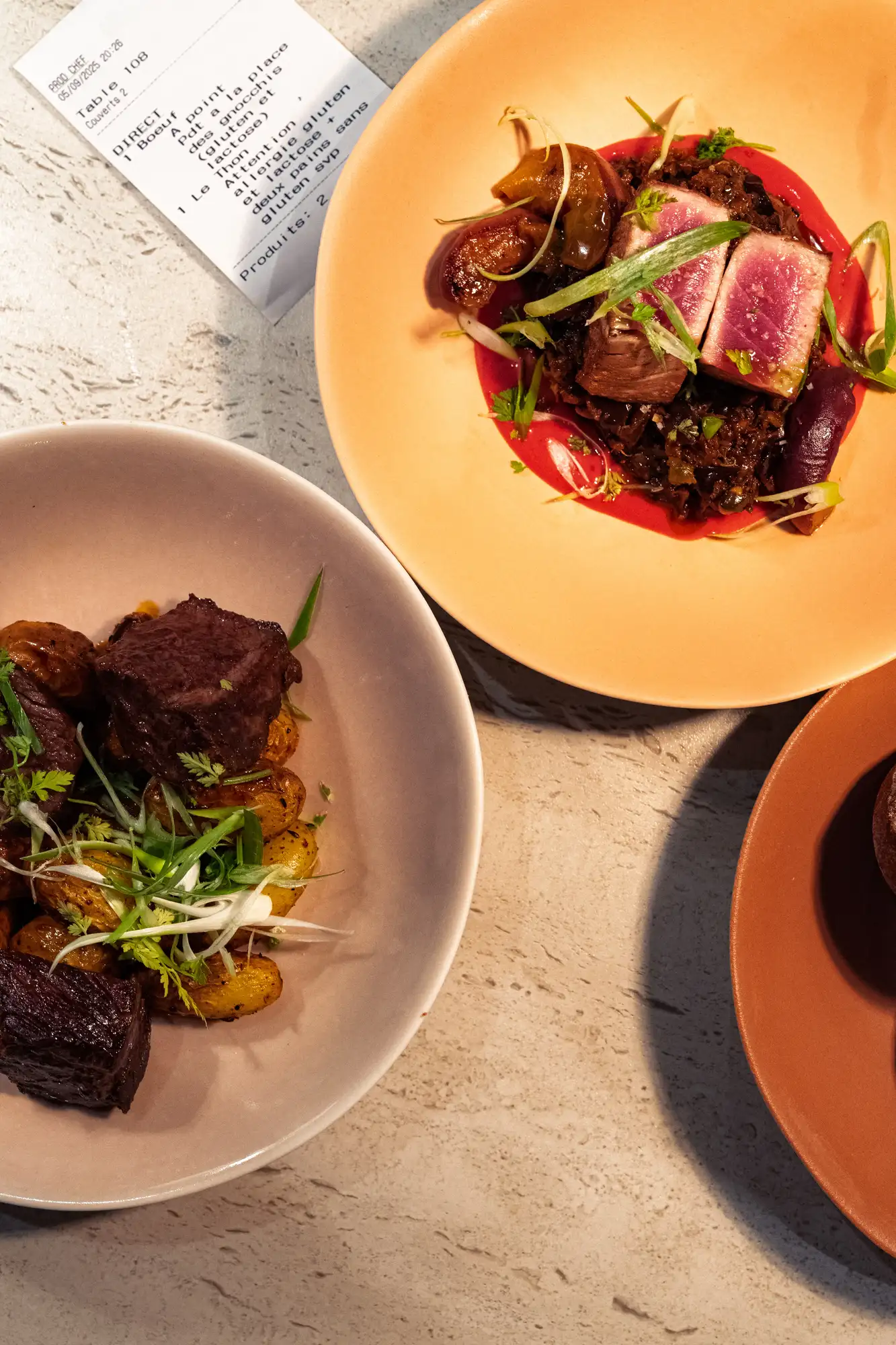 Deux assiettes contenant des plats raffinés : une avec des morceaux de viande saignante sur une purée rouge avec des herbes, l'autre avec des morceaux de viande braisée sur des pommes de terre rôties garnies de fines herbes, posées sur une table claire près d'un reçu de commande.