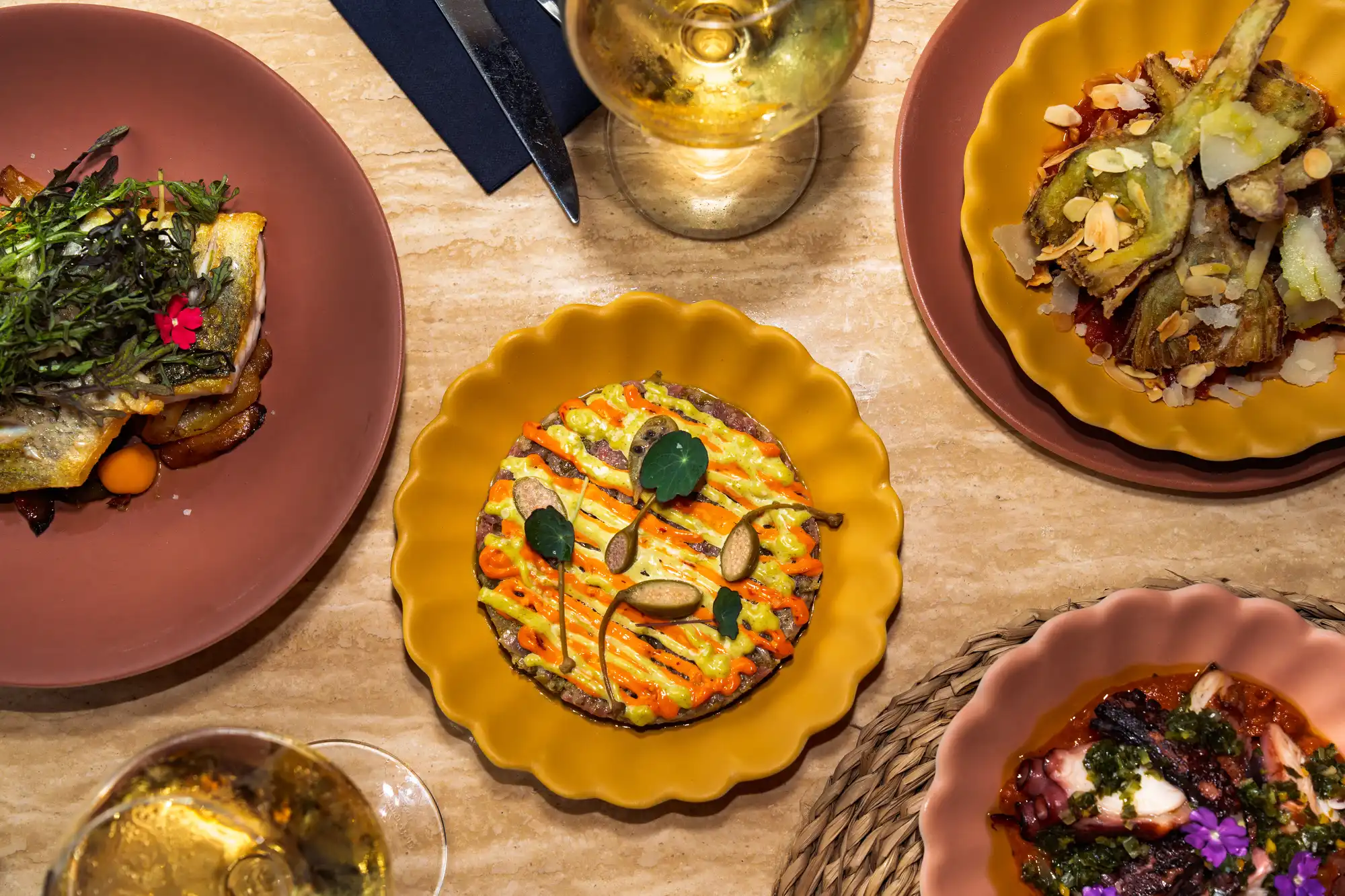 Quatre plats gastronomiques colorés sur une table, comprenant un plat central rond avec sauces orange et jaune, entouré de plats contenant du poisson avec herbes, des artichauts grillés et une préparation de poulpe avec fleurs comestibles, accompagnés de deux verres de vin blanc.