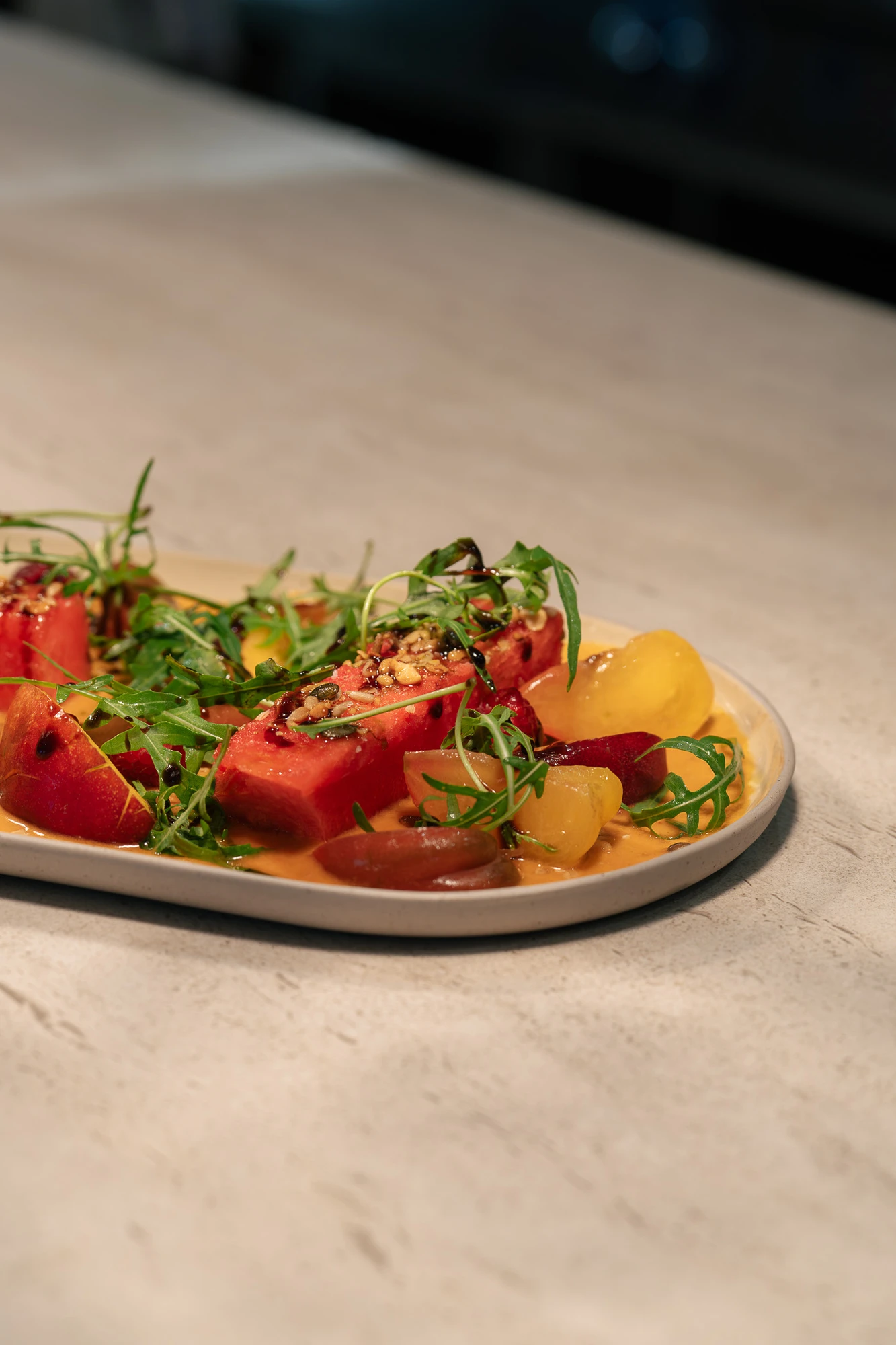 Assiette contenant des morceaux de pastèque, tomates cerises jaunes et rouges, roquette, et une vinaigrette avec des graines sur le dessus, posée sur une surface en pierre claire.