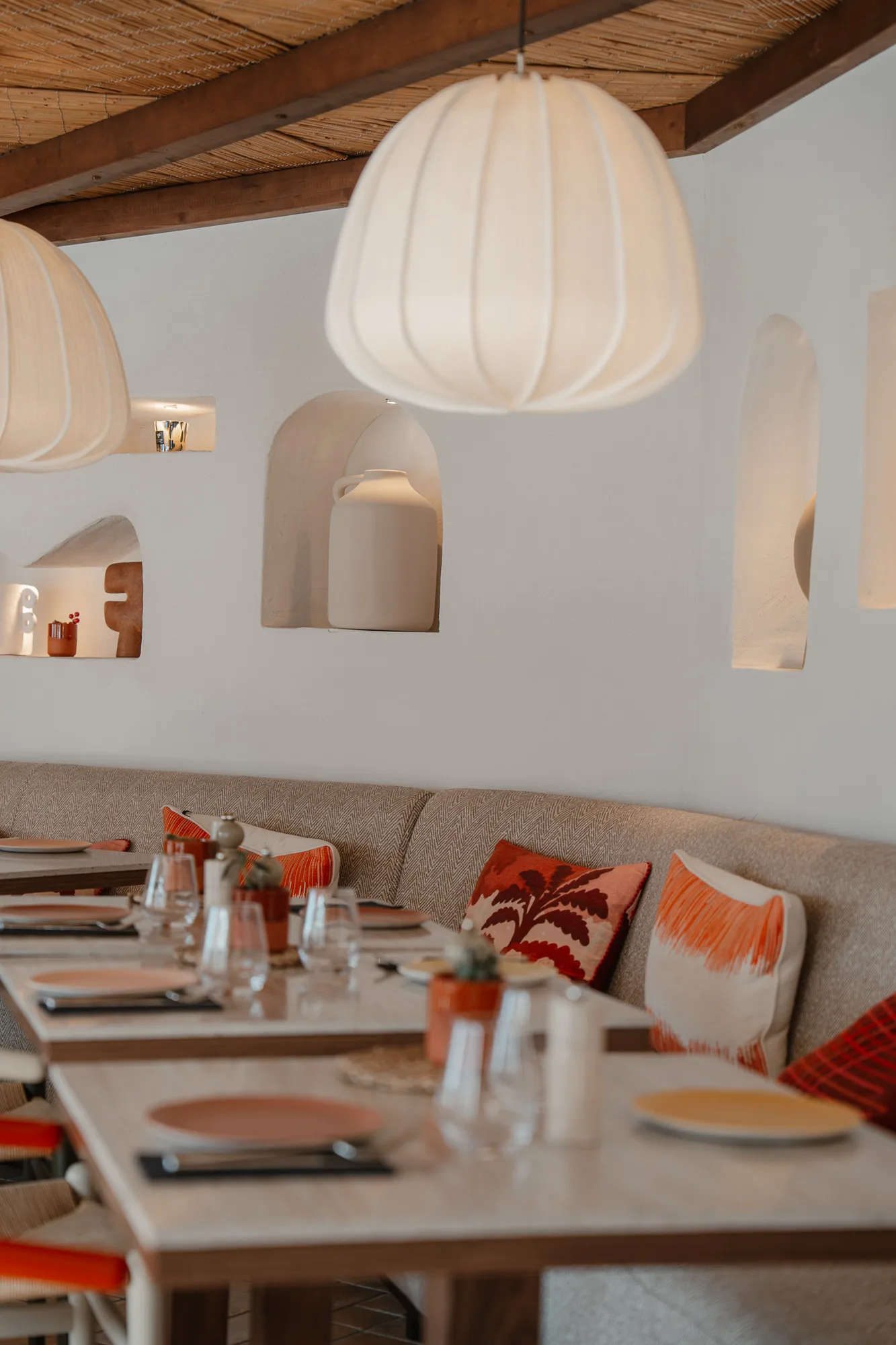 Intérieur d'un restaurant avec tables dressées de verres, assiettes et couverts, banquette beige avec coussins rouges et oranges, lampes suspendues blanches et décorations murales en poterie.