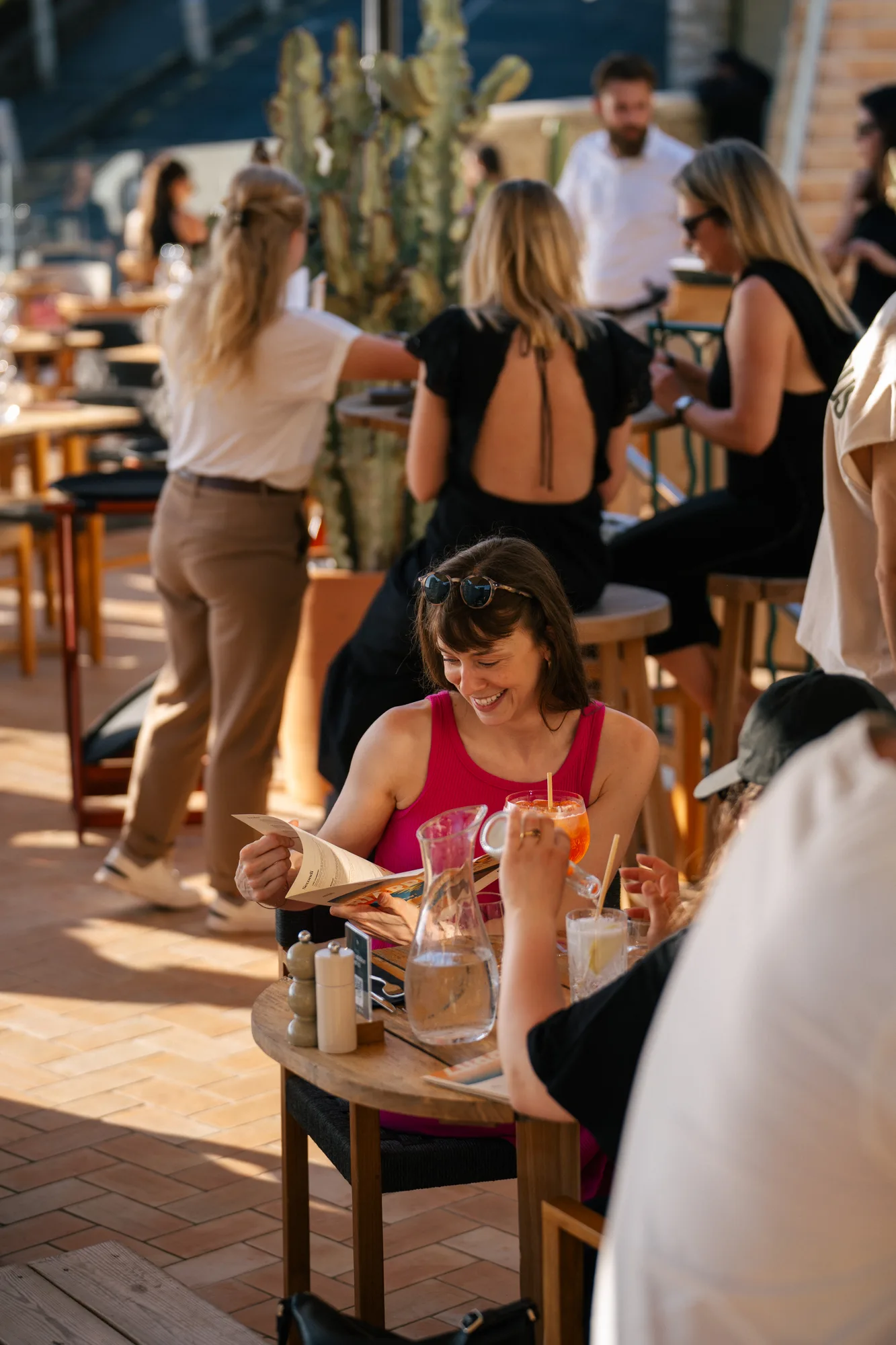 Une femme souriante en débardeur rose assise à une table ronde en bois dans un café en plein air, tenant un menu, avec une carafe d'eau et un verre à boisson orange sur la table, entourée d'autres personnes.