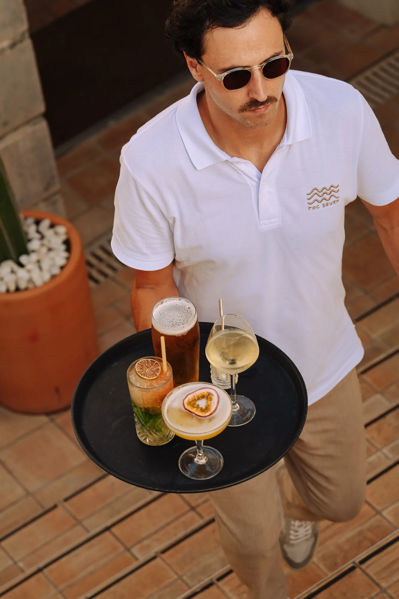 Homme portant des lunettes de soleil et un polo blanc avec logo, tenant un plateau noir avec quatre verres de boissons variées, dont une bière, un cocktail avec tranche de fruit sec, un verre de vin blanc et un cocktail avec fruit de la passion en garniture.