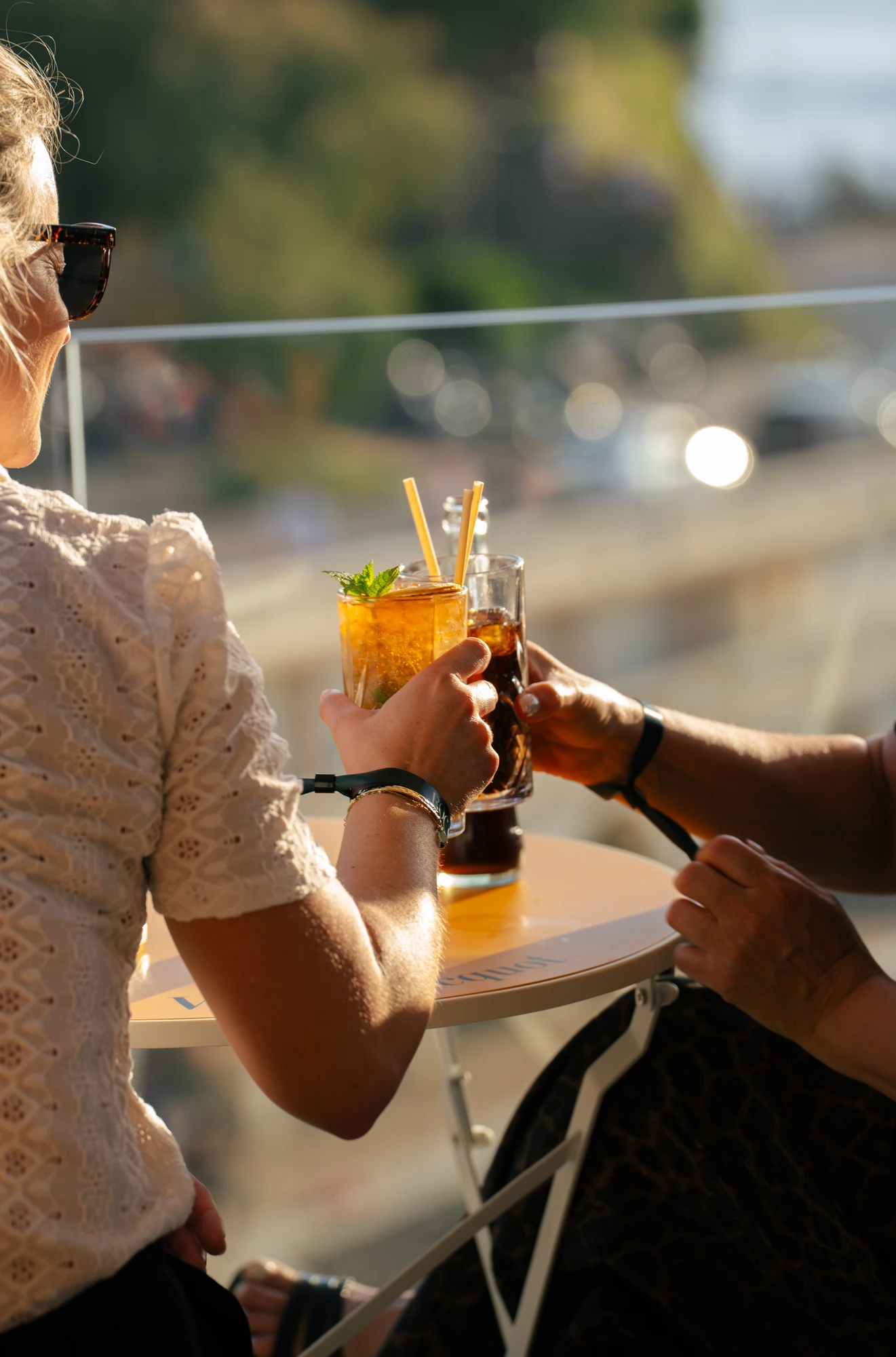 Deux personnes tenant des verres de boissons rafraîchissantes en train de trinquer à une petite table ronde en extérieur sous une lumière chaude.