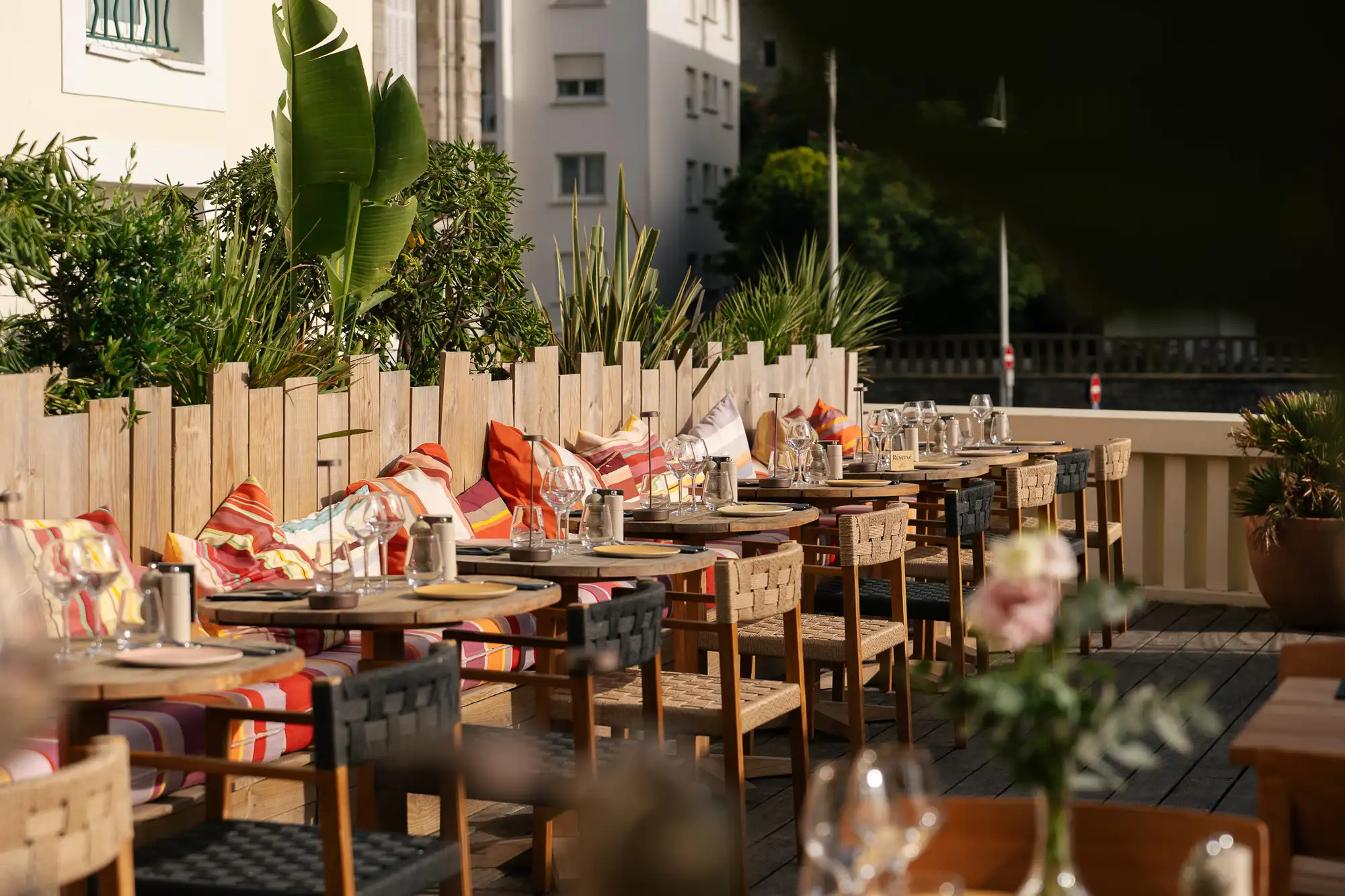 Terrasse extérieure d'un restaurant avec tables en bois dressées avec assiettes, verres et serviettes, chaises tissées et coussins colorés rayés le long d'une palissade en bois, entourée de plantes vertes.