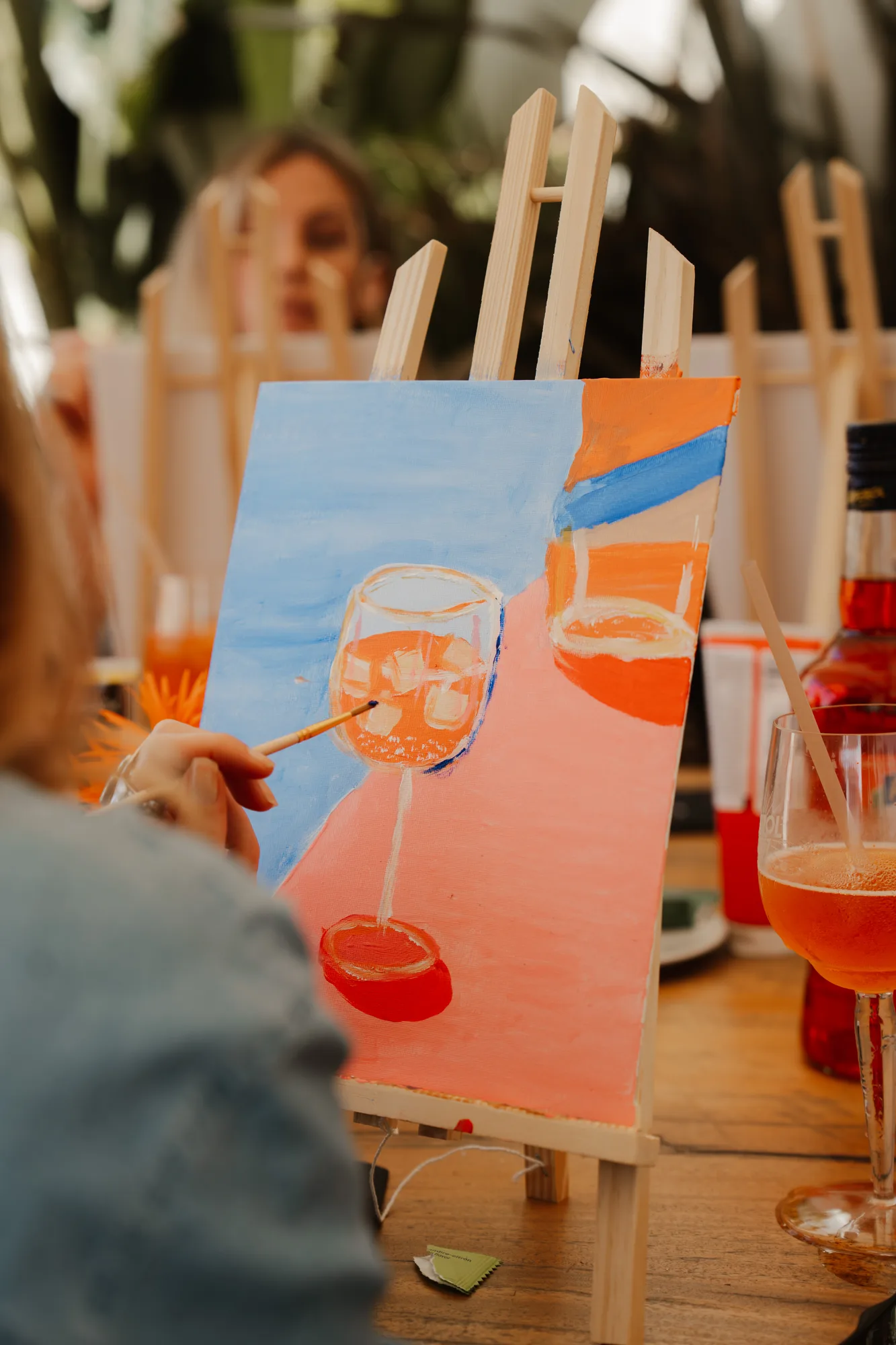 Person peint une toile sur un chevalet représentant un verre à vin avec des glaçons sur un fond aux couleurs bleu et rose, avec un verre à cocktail orange posé à côté sur une table en bois.