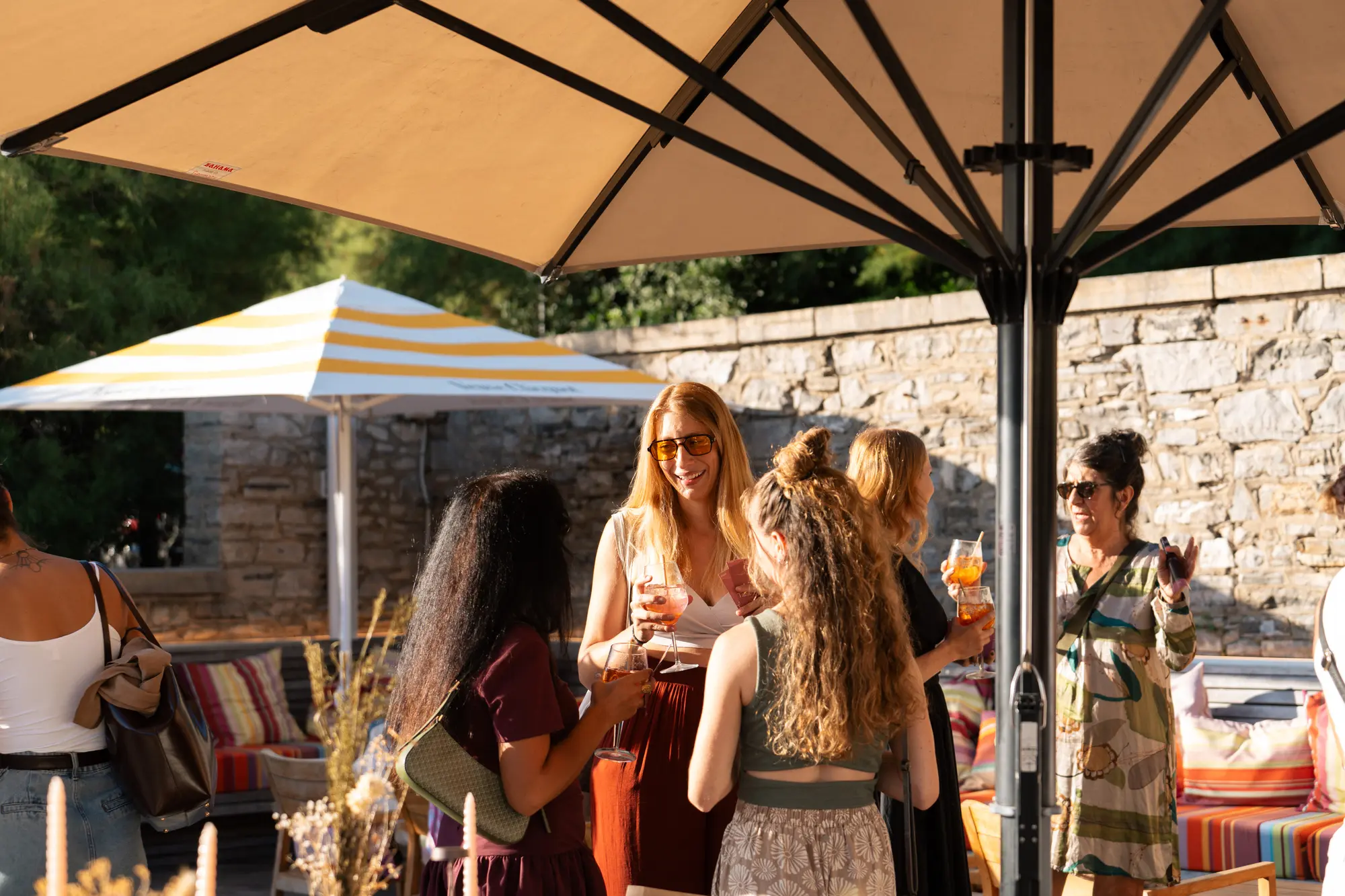 Groupe de femmes debout sous un grand parasol extérieur, discutant et tenant des verres avec des boissons orange, décor estival avec coussins rayés colorés et mur en pierre en arrière-plan.