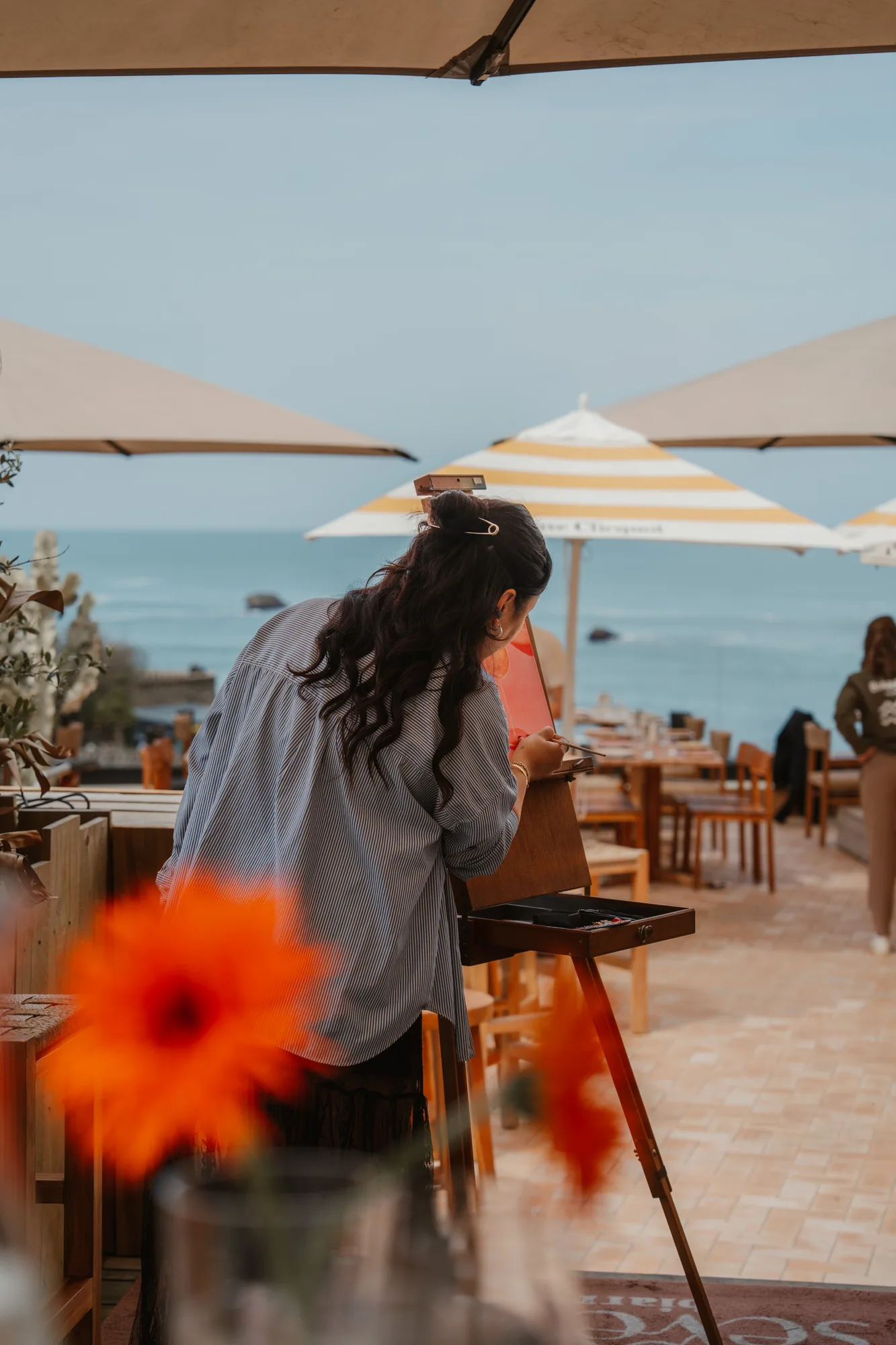 Une femme est penchée devant un chevalet de peinture en bois sur une terrasse extérieure avec vue sur la mer, des parasols blancs et jaunes, et des tables en bois autour, une fleur orange floue en premier plan.