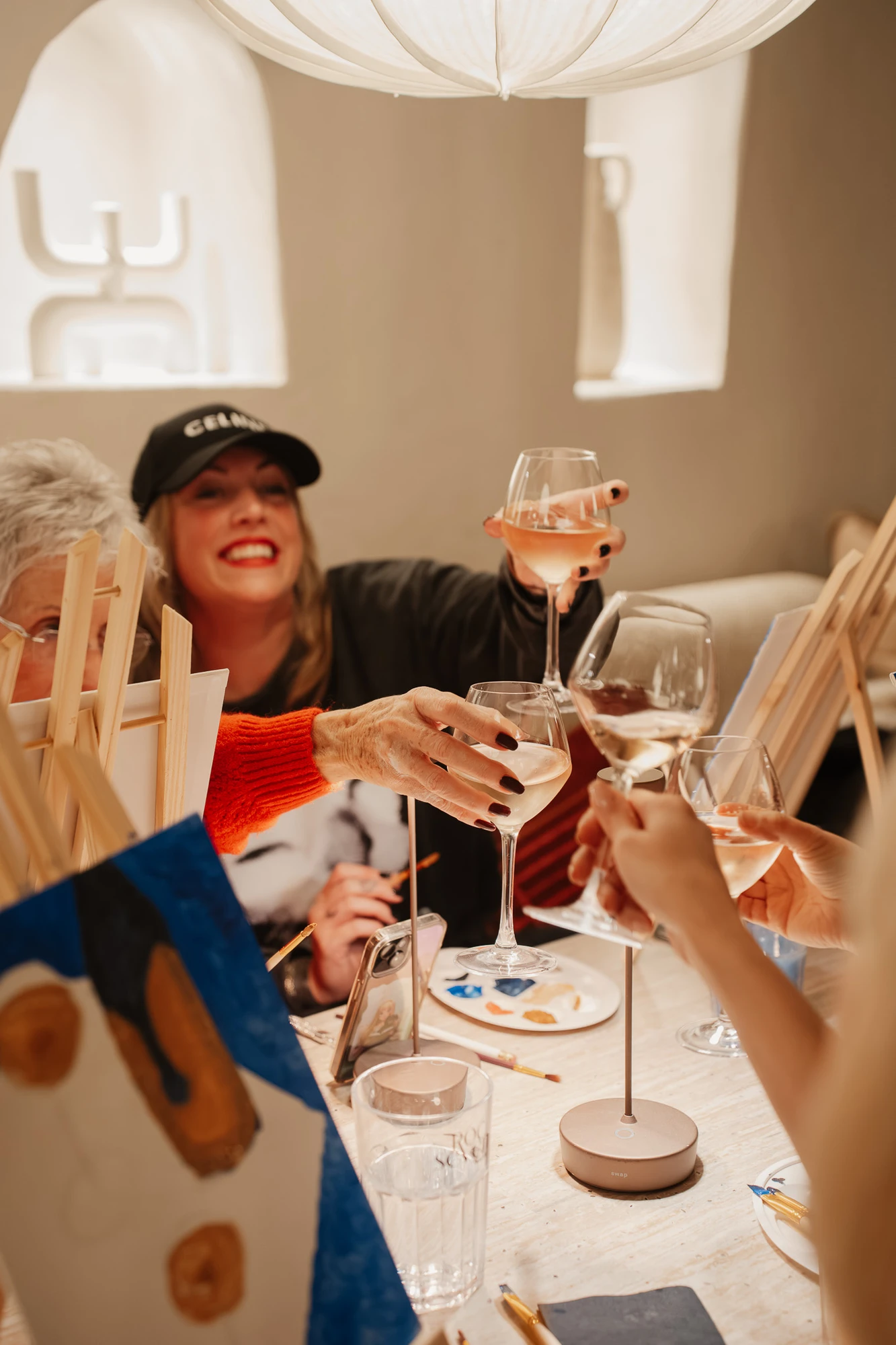 Des femmes trinquent avec des verres de vin rosé autour d'une table où des chevalets et des pinceaux sont posés, suggérant une soirée peinture et vin conviviale.