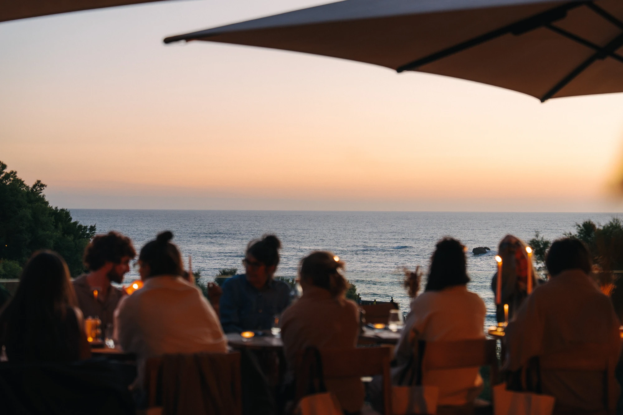 Un groupe de personnes est assis à une table en plein air sous un parasol, avec des bougies allumées, regardant la mer au coucher du soleil.