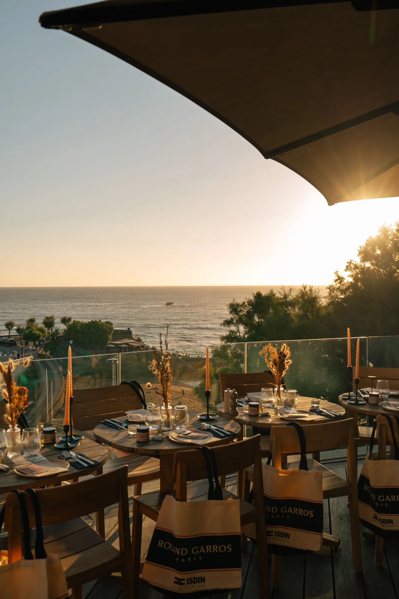 Terrasse de restaurant avec tables en bois dressées, décorées de bougies et de bouquets de fleurs séchées, vue sur la mer au coucher du soleil, avec des sacs Roland Garros suspendus aux chaises.