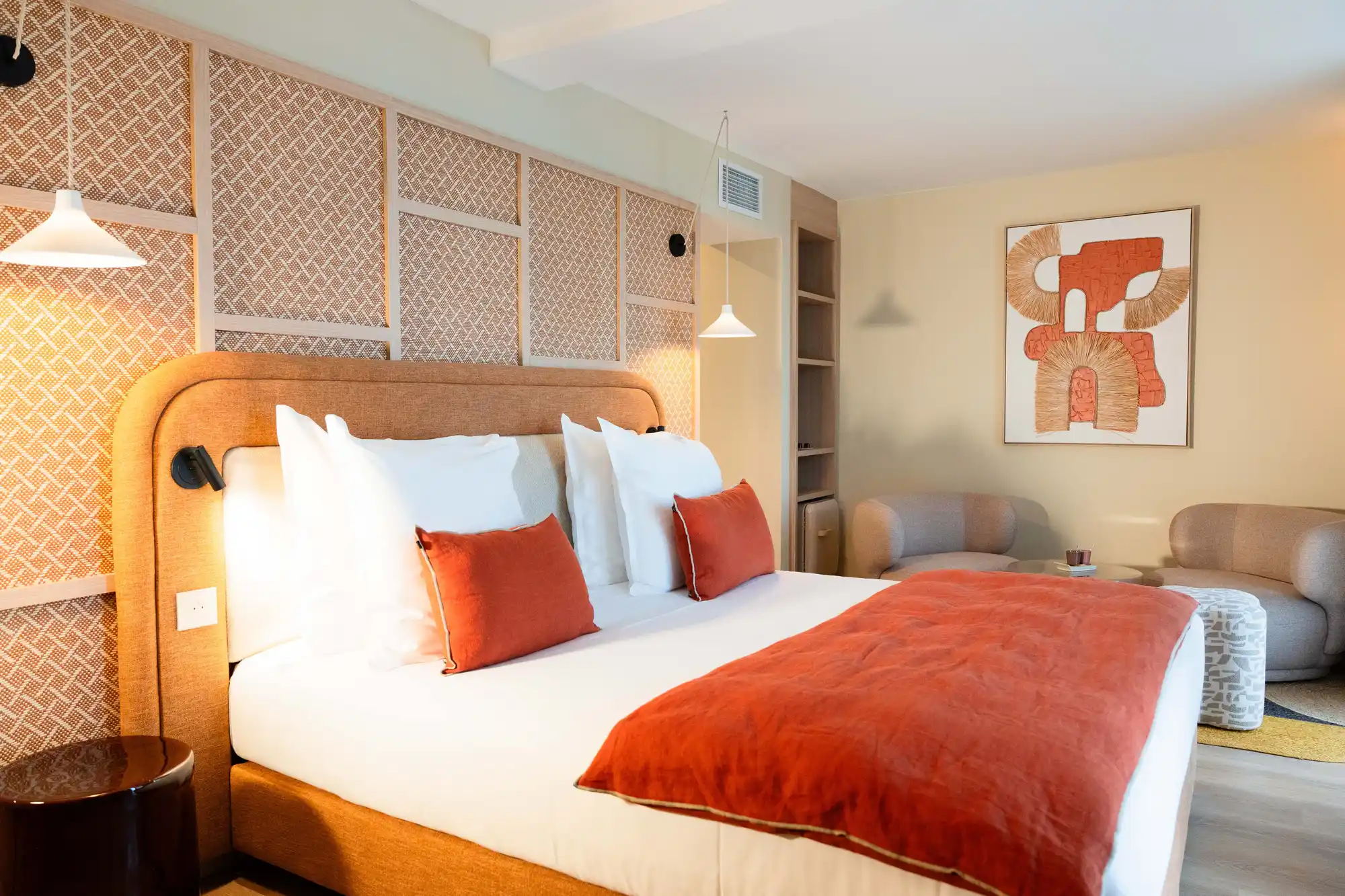 Chambre moderne avec grand lit double aux coussins et couverture orange, lampes suspendues blanches, murs clairs avec panneau en tissu tissé derrière le lit, deux fauteuils beige arrondis avec petite table basse, et tableau abstrait aux tons orange et beige.