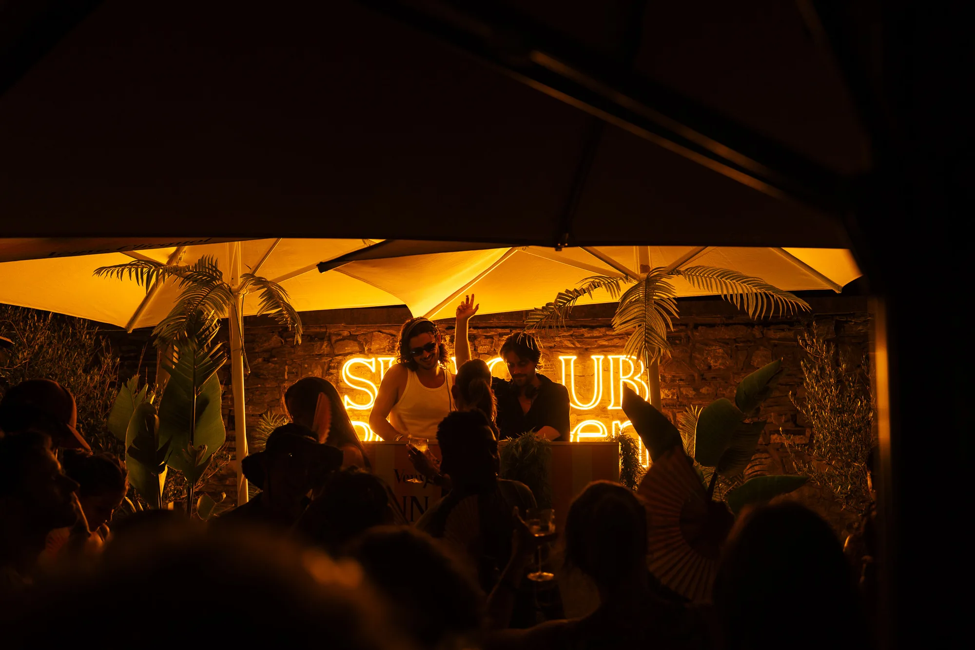 Deux DJ souriants jouent de la musique sous des parasols éclairés par une enseigne lumineuse orange, entourés de plantes et d'une foule en fête.