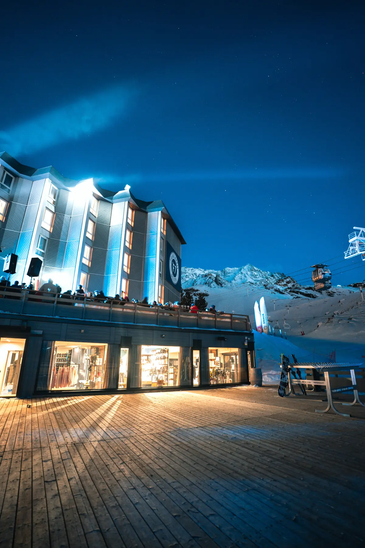 Bâtiment éclairé d'une station de ski en soirée avec terrasse, skieurs sur la terrasse, montagnes enneigées et télécabines visibles sous un ciel étoilé.