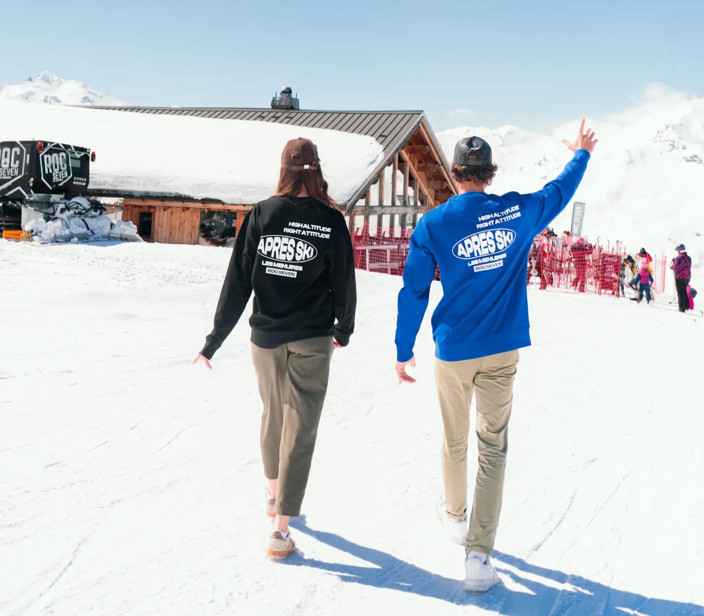 Deux personnes marchant sur la neige vers un chalet en montagne, portant des pulls avec le texte "APRES SKI LES MENUIRES ROC SEVEN" vus de dos, l'une en bleu et l'autre en noir, avec un ciel clair et des montagnes enneigées en arrière-plan.