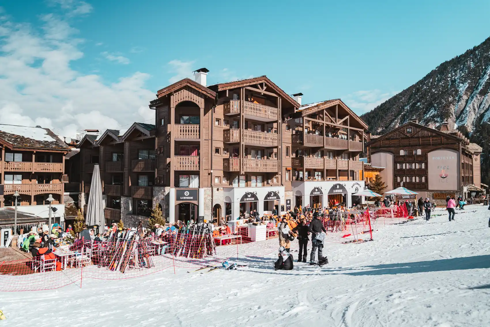 Station de ski animée avec des groupes de personnes en tenue de ski autour d'un bâtiment en bois avec balcons, entouré de montagnes enneigées par une journée ensoleillée.