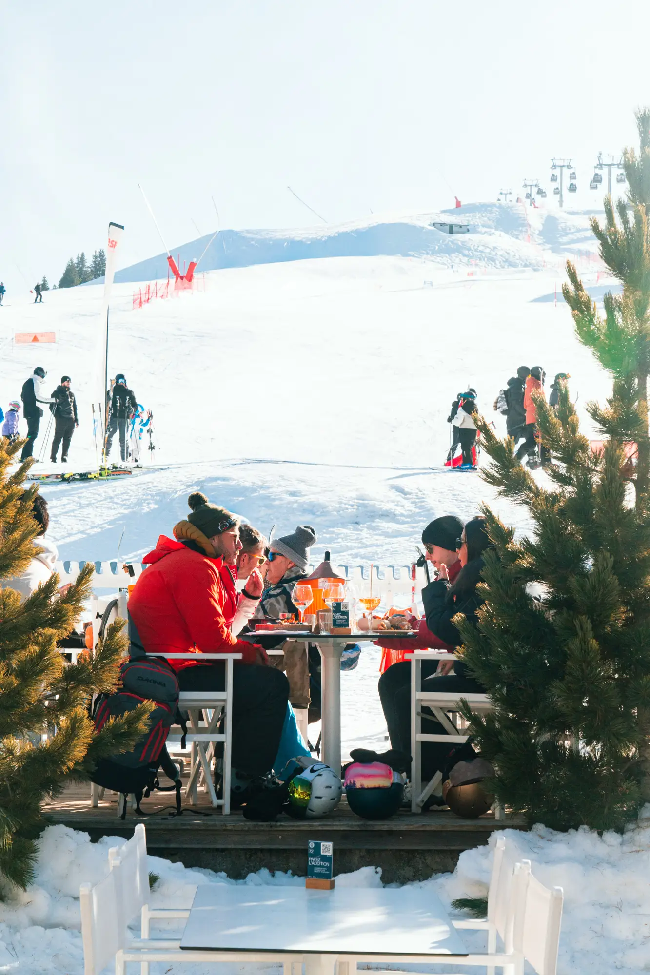 Groupe de personnes assises autour d'une table extérieure aux sports d'hiver, avec des casques et des boissons, sur fond de piste de ski et télésièges.