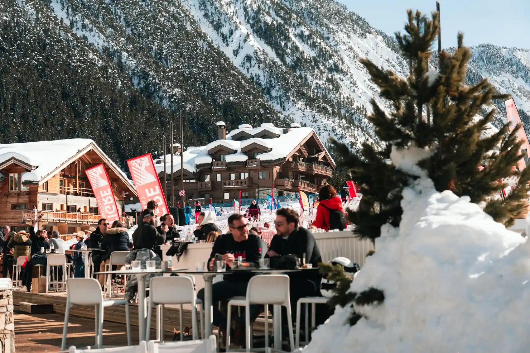 Des personnes profitent d'une terrasse ensoleillée dans un village de montagne enneigé avec des chalets en bois et une grande montagne en arrière-plan.
