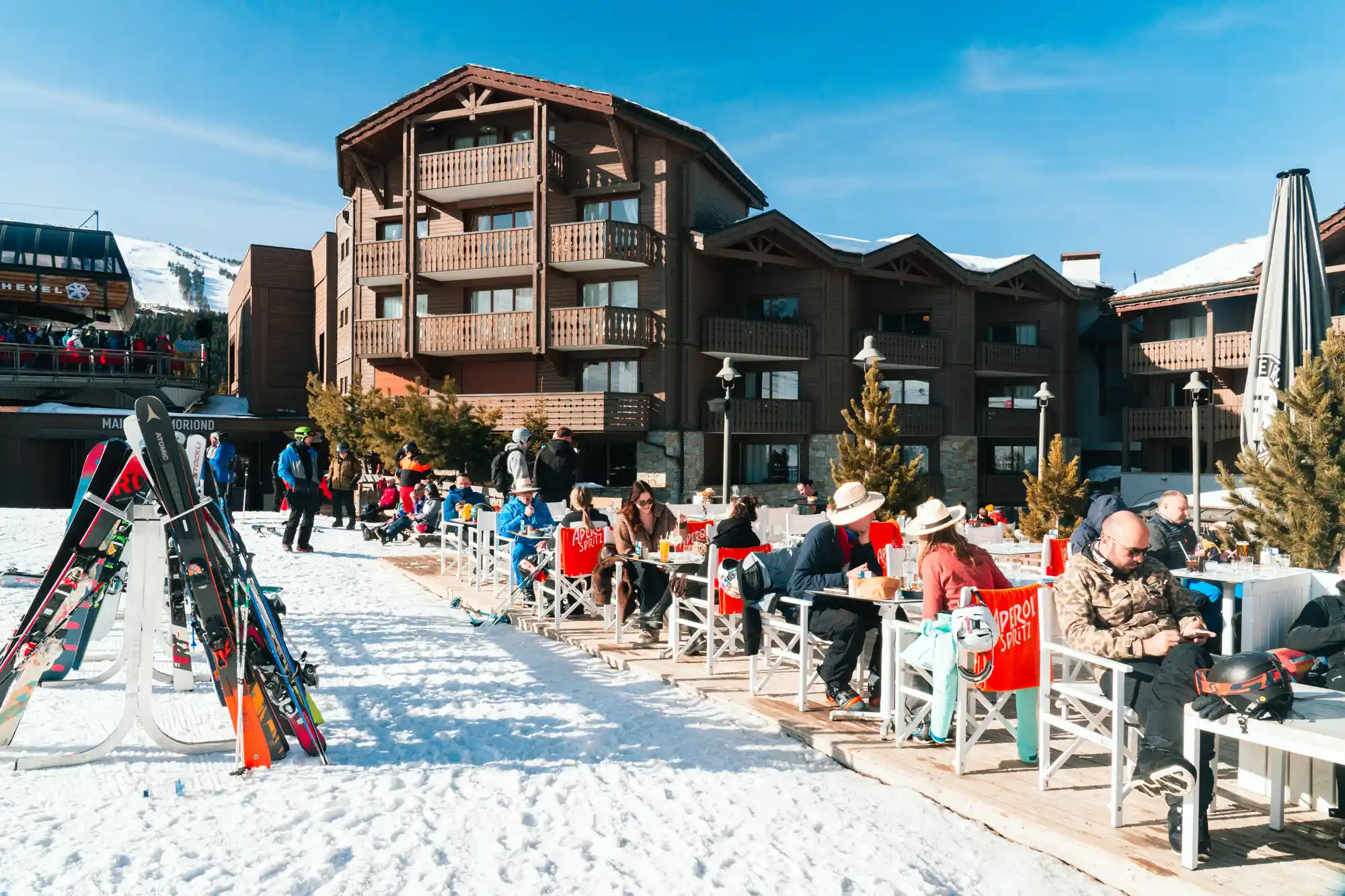 Terrasse extérieure d'un restaurant en station de ski avec plusieurs personnes assises en train de manger et boire, entourées de neige, au pied d'un grand bâtiment en bois et sous un ciel bleu clair.