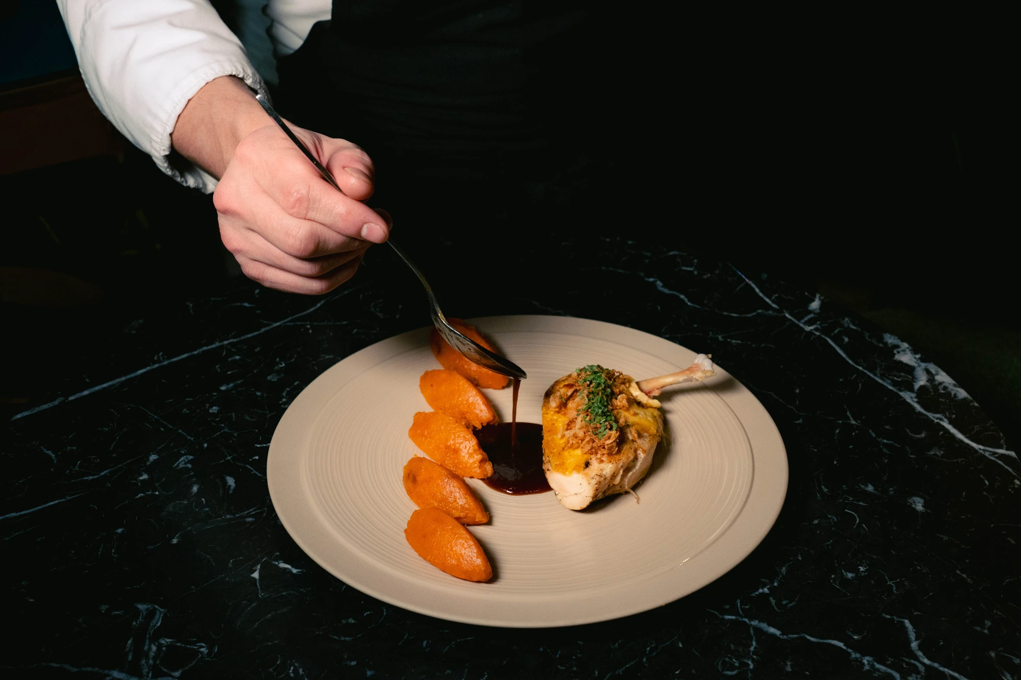 Une main verse une sauce foncée à l'aide d'une cuillère sur une assiette avec un morceau de poulet rôti garni d'herbes et plusieurs quenelles de purée orange, posée sur une surface noire marbrée.