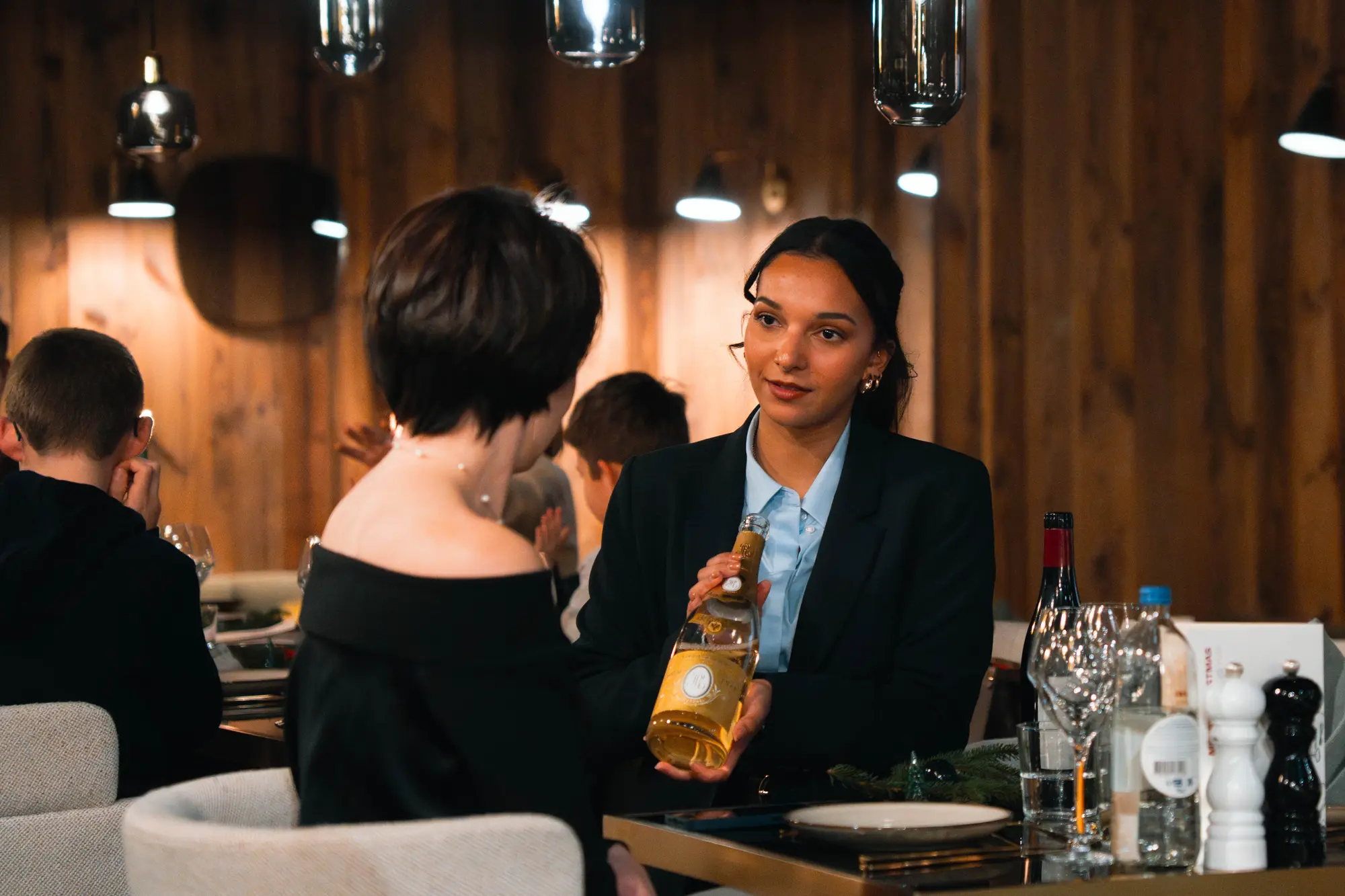 Une serveuse vêtue d'un blazer noir présente une bouteille de vin blanc à une cliente en robe noire assise à une table dans un restaurant au décor en bois avec plusieurs verres et bouteilles sur la table.