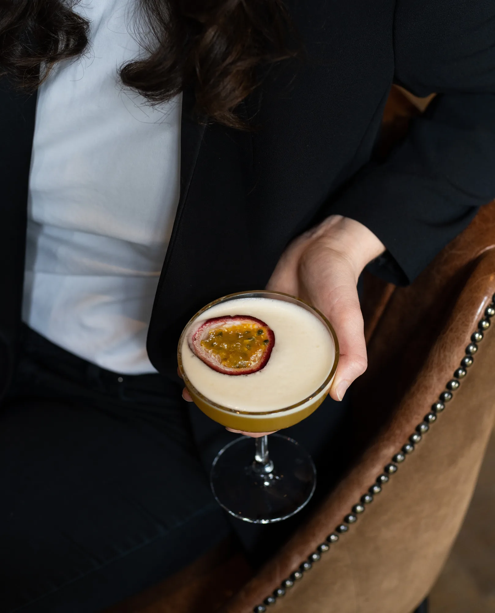 Person assise tenant un verre à pied avec un cocktail jaune surmonté de mousse blanche et une tranche de fruit de la passion.