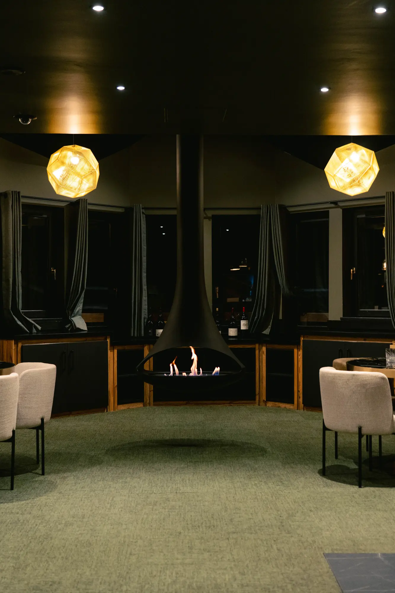 Intérieur d'un salon moderne avec cheminée suspendue allumée au centre, deux suspensions lumineuses géométriques jaunes, fauteuils beiges et rideaux gris devant les fenêtres.