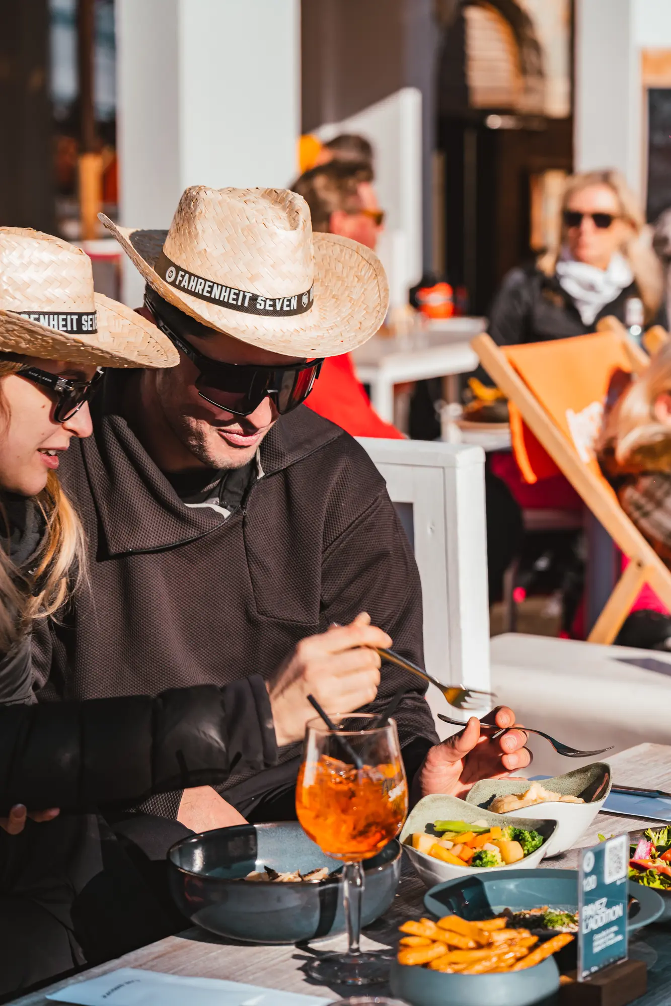 Deux personnes portant des chapeaux de paille avec l'inscription Fahrenheit Seven mangent ensemble à une table en plein air avec des plats colorés et une boisson orange.