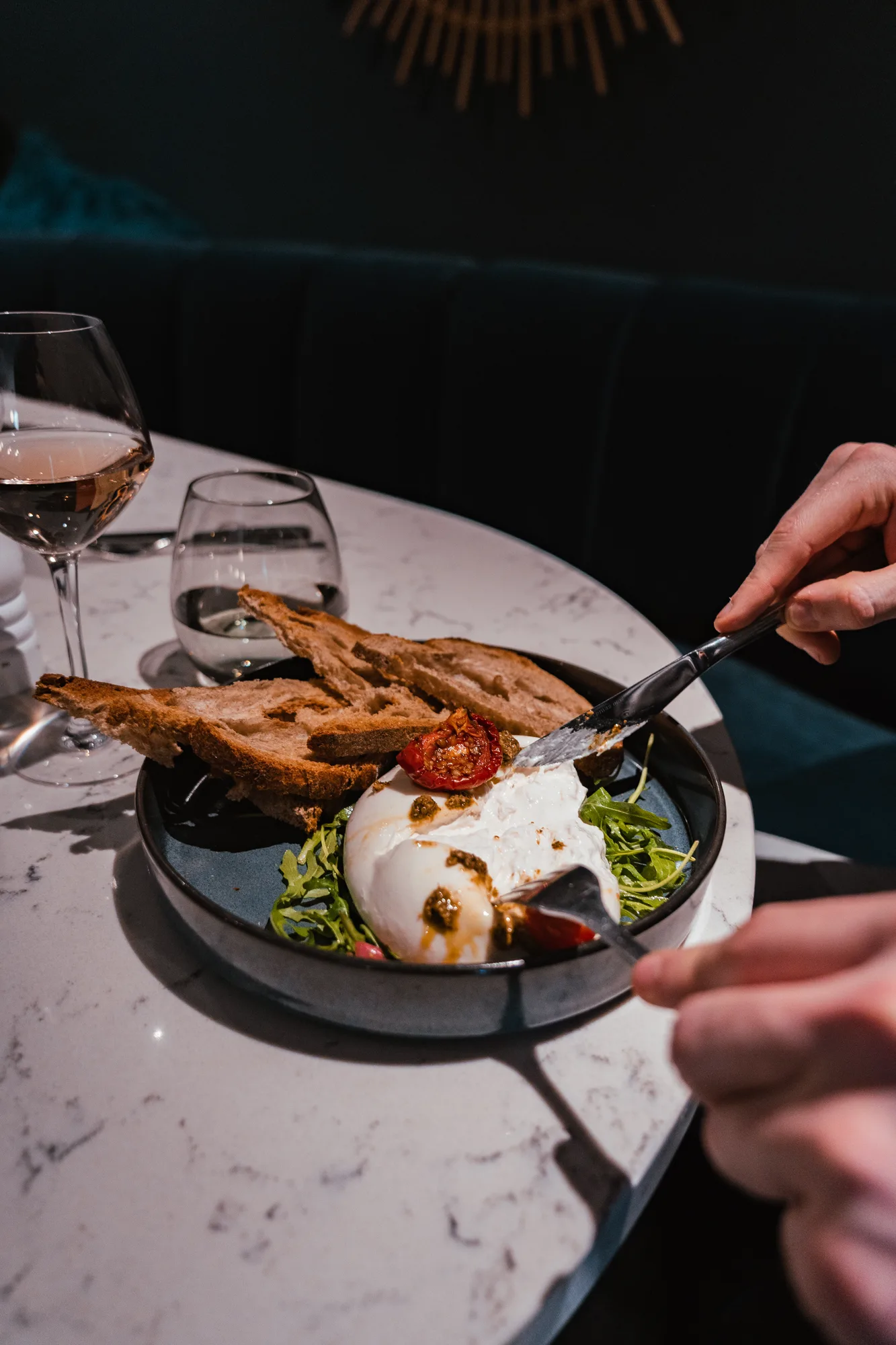 Person coupant une burrata crémeuse sur un lit de roquette avec du pain grillé et une tomate séchée dans une assiette noire, sur une table en marbre avec un verre de vin rosé et un verre d'eau à côté.