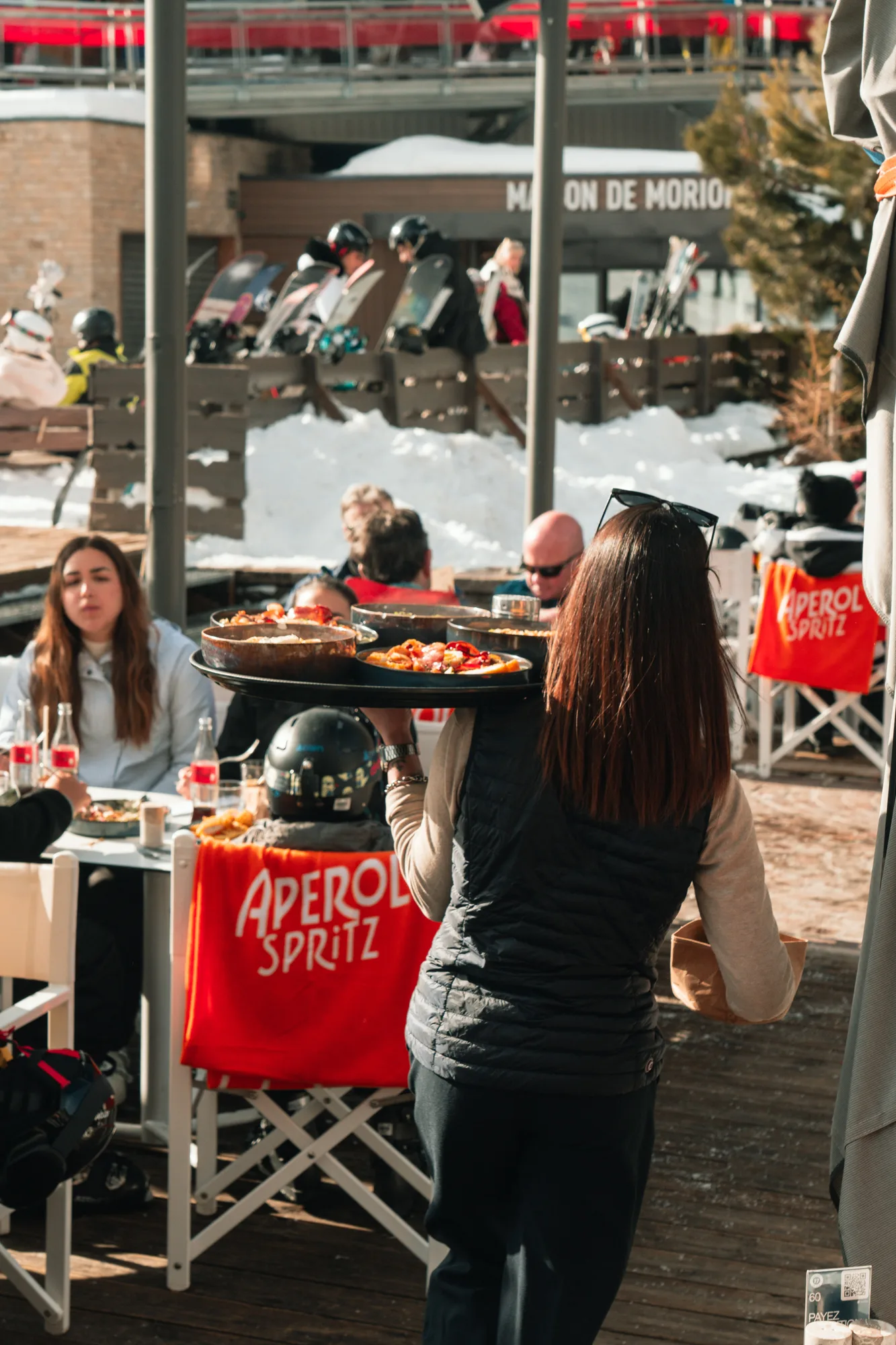 Serveuse portant un plateau avec plusieurs plats dans un restaurant en plein air d'une station de ski enneigée avec des clients assis autour de tables et des casques de ski posés sur des chaises avec couvertures Aperol Spritz.