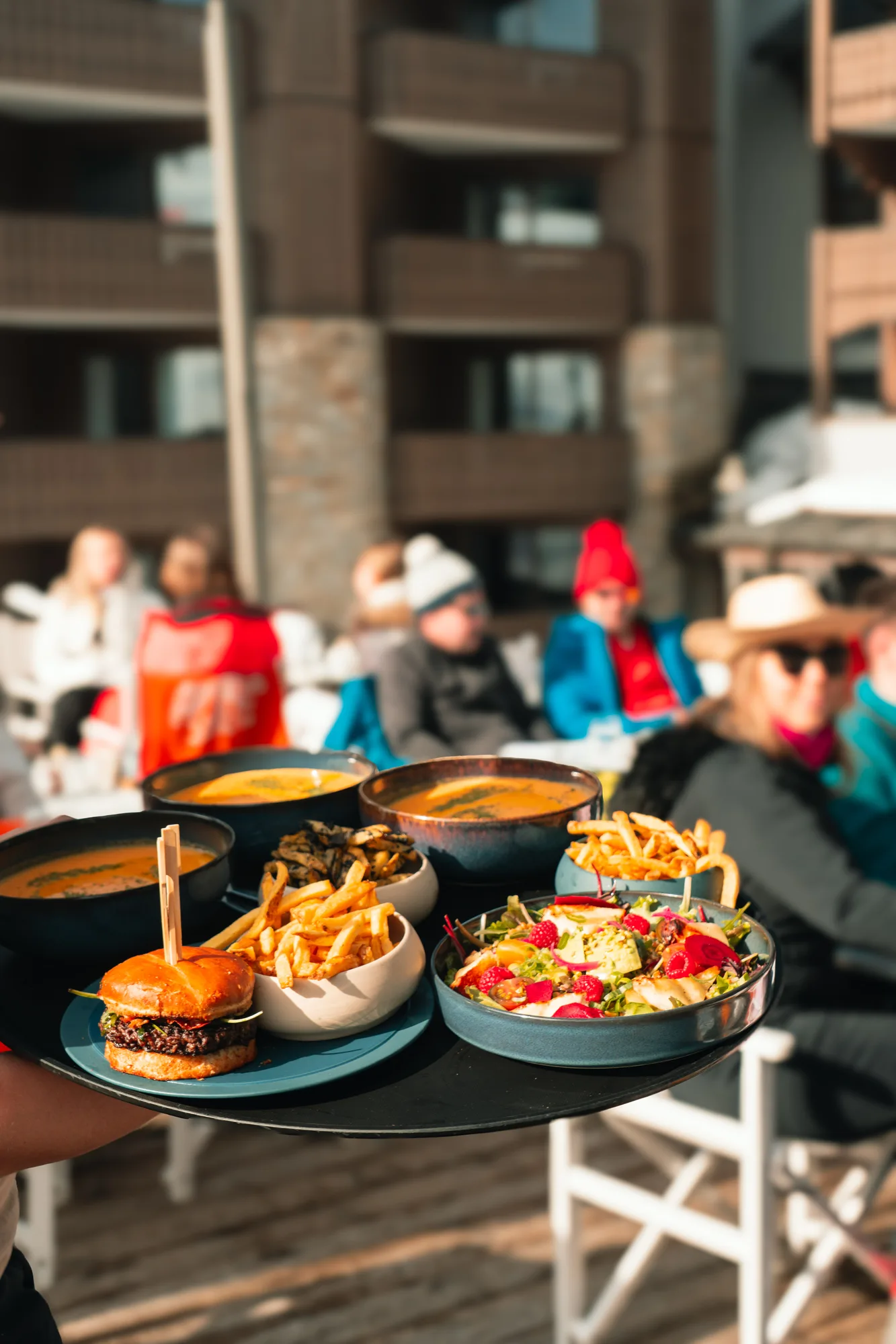 Un plateau avec un hamburger, des frites, une salade avec des framboises, et plusieurs bols de soupe, porté vers des personnes habillées chaudement en extérieur.