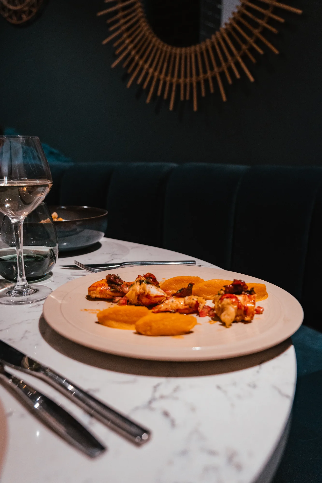Assiette blanche sur une table en marbre avec un plat gastronomique composé de morceaux de viande ou de poisson grillé et des quenelles de purée orange, accompagné d’un verre de vin blanc et d’un verre d’eau dans un cadre élégant avec banquette sombre et décoration murale en bois.